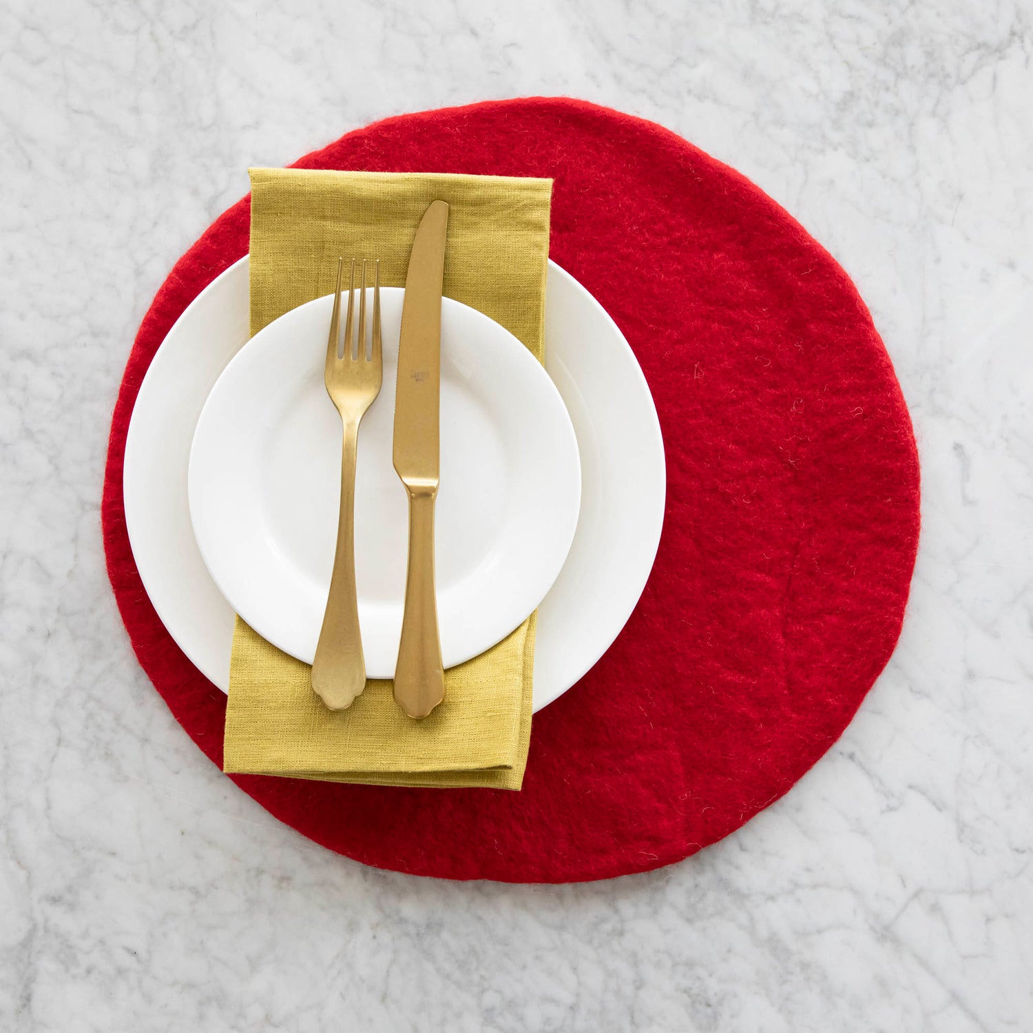A place setting with the Red Felt Placemat, white dinner plates with a gold napkin in between and a gold fork and knife atop, on a marble table.