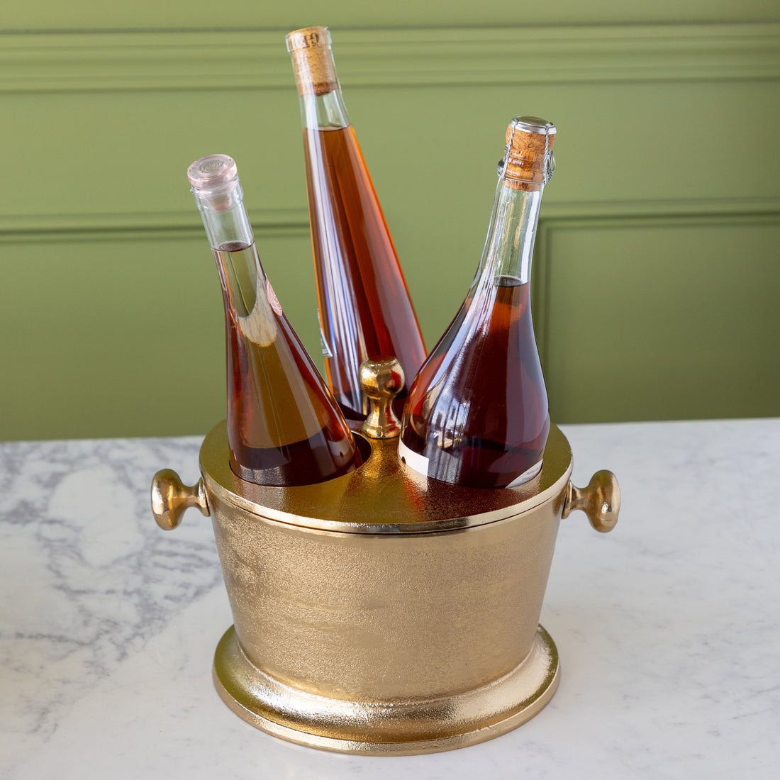 Three bottles of alcohol in the Gold Raw Aluminum Bottle Holder against a green wall.