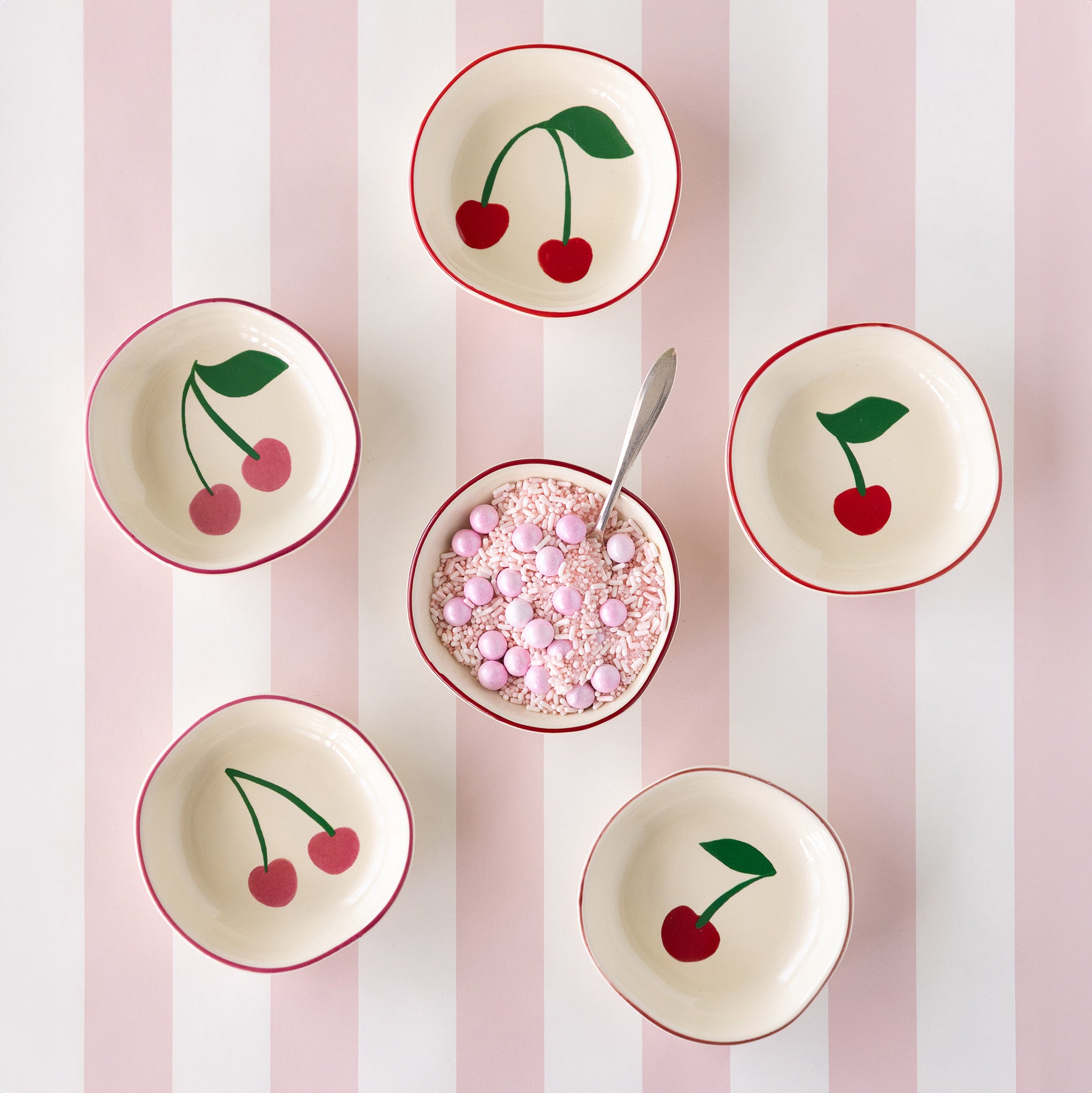 Six Very Cherry pinch bowls sit on a Pink Classic Stripe Runner; five show off their cherry designs, while the center bowl holds pink sprinkles and candy and a spoon.