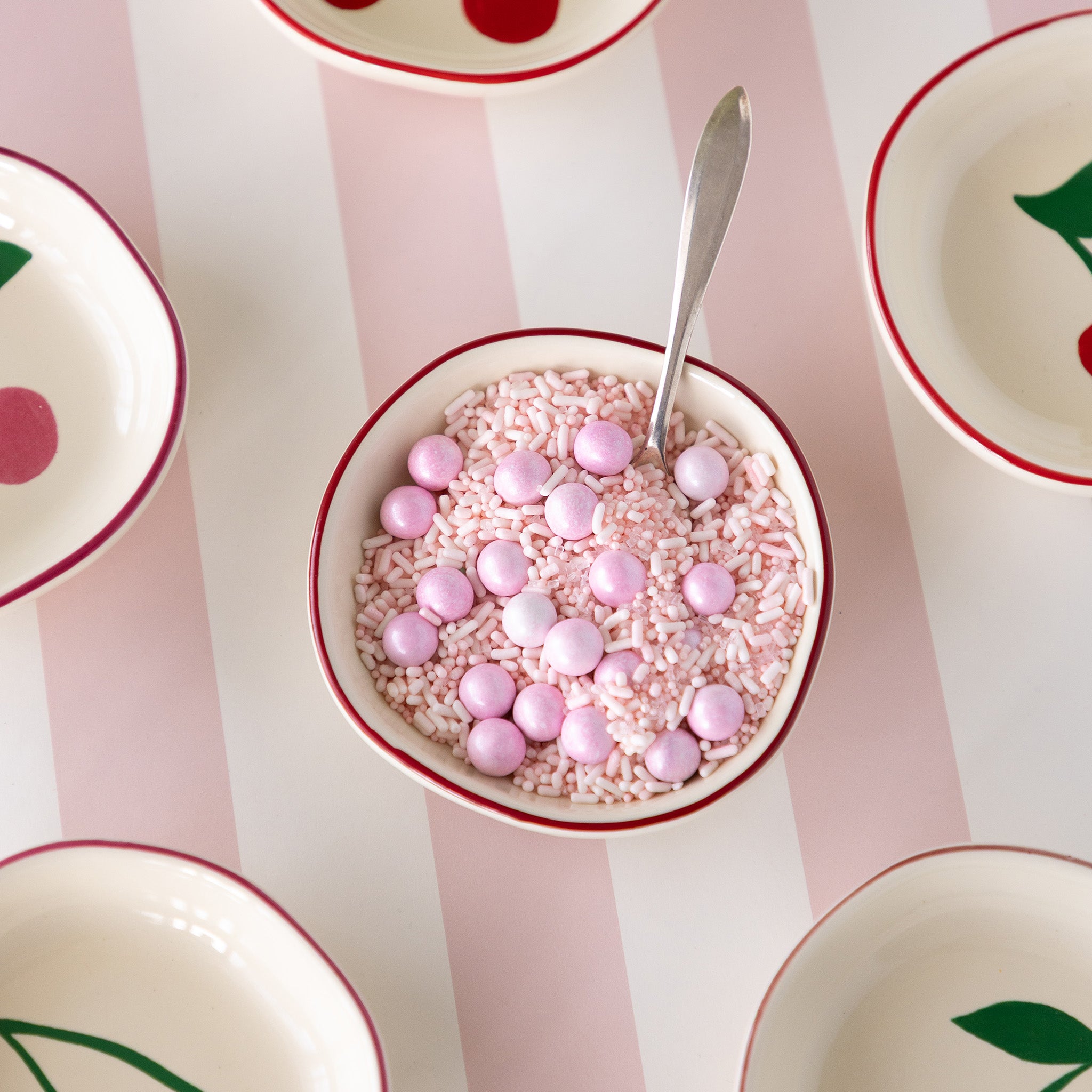 A Very Cherry Pinch bowl filled with pink sprinkles and round balls, and a spoon.