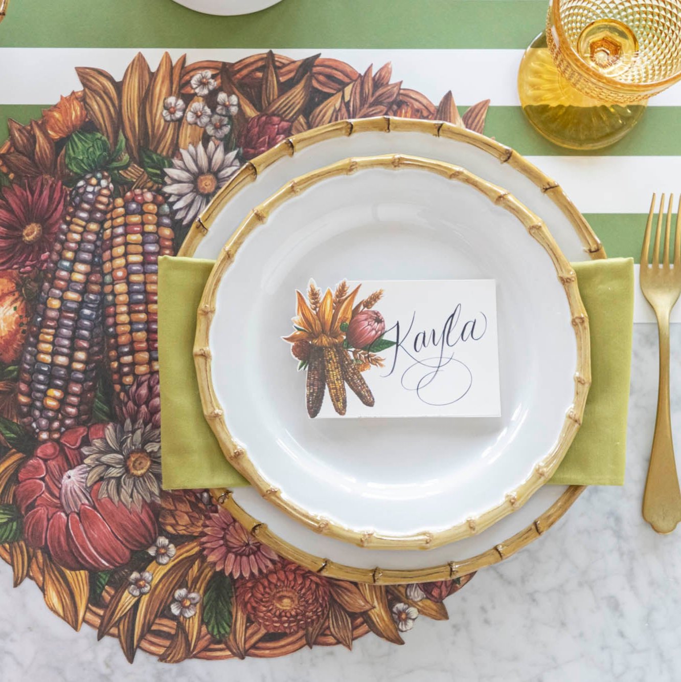 An elegant table setting featuring the Gathering Place Card with "Kayla" written on it, resting on bamboo dinnerware, on top of the Die-cut Gathering Basket Placemat.