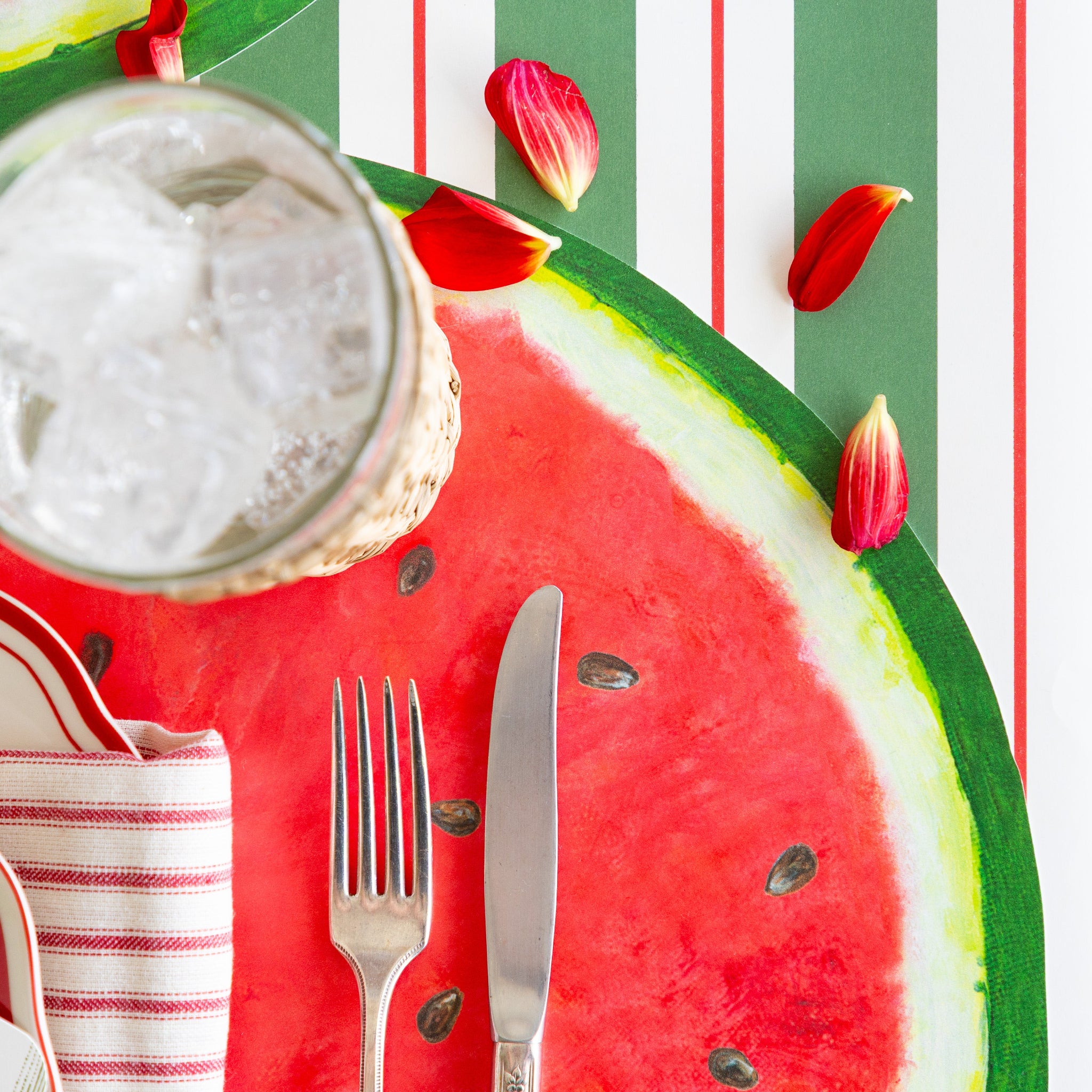 Place setting with the Die-cut Watermelon Placemat, glass of water, silverware and flower petals on the Green & Red Awning Stripe Runner.