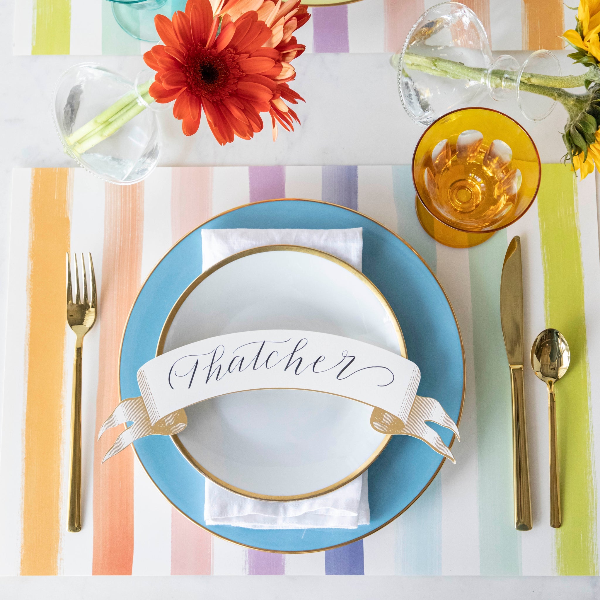 Colorful table setting with the Sorbet Painted Stripe Placemat, plates with the Gold Banner Table Accent atop, gold flatware and vases of flowers.