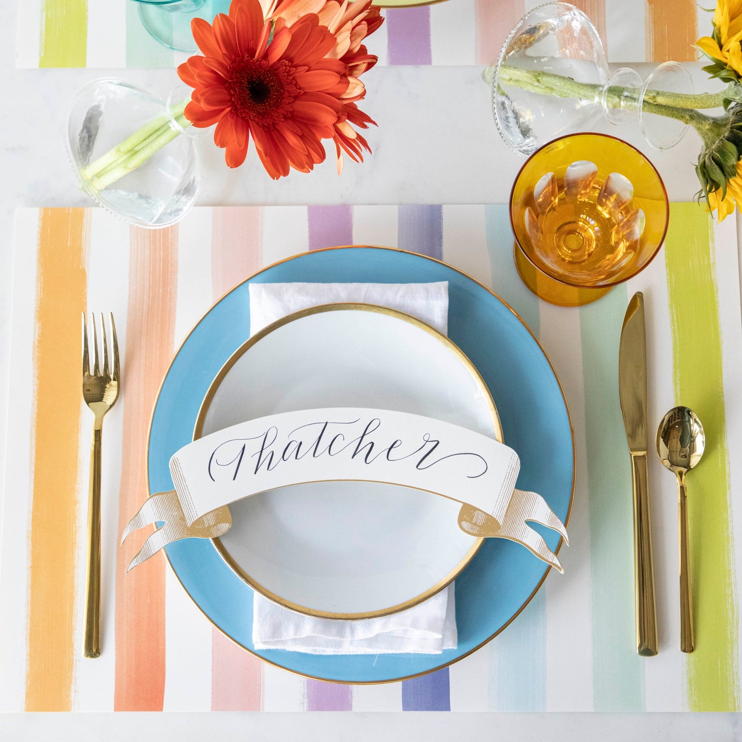 Colorful table setting with the Sorbet Painted Stripe Placemat, plates with the Gold Banner Table Accent atop, gold flatware and vases of flowers.