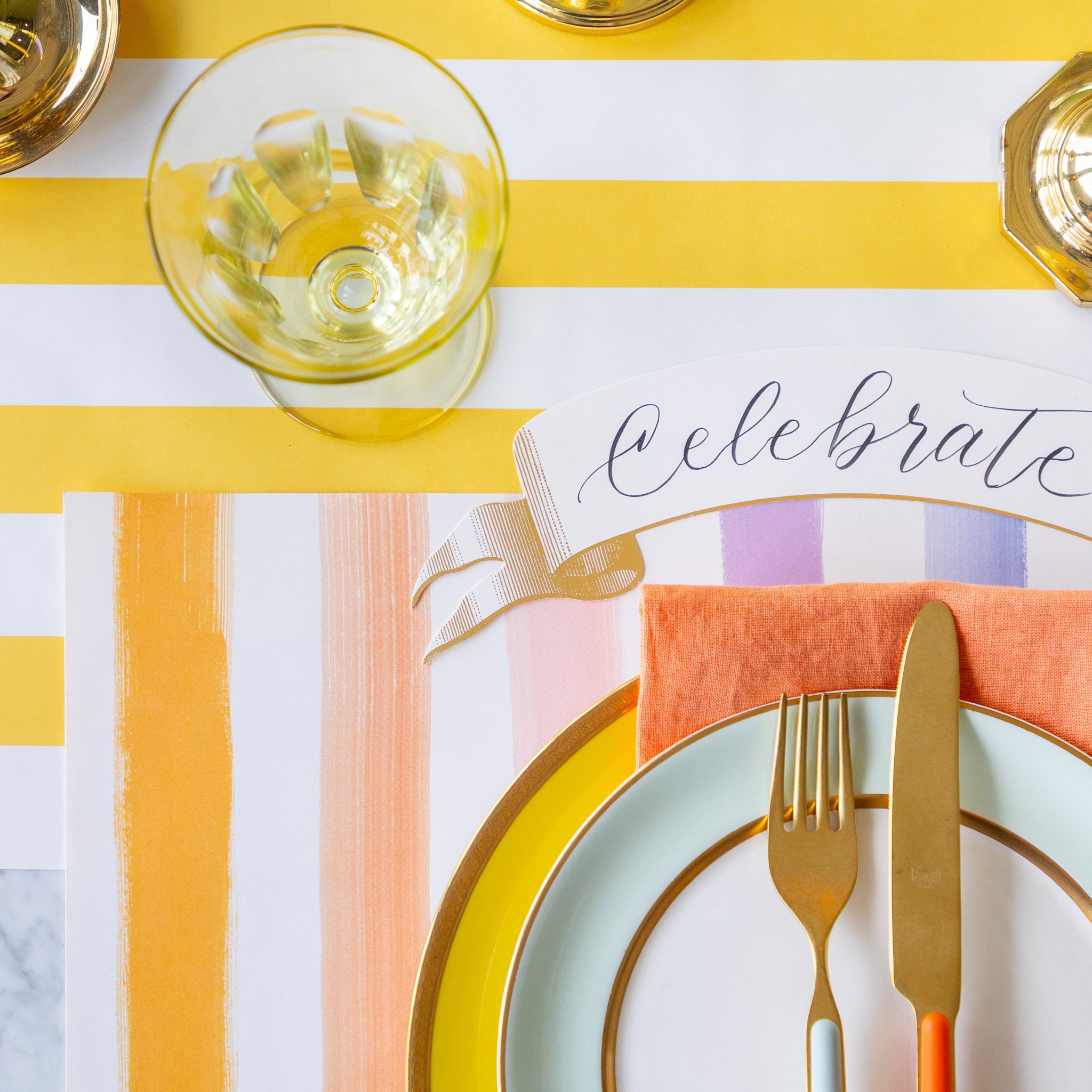 Colorful table setting with the Sorbet Painted Stripe Placemat, colorful plates and cutlery atop, and Classic Gold Banner Table Accent on the Marigold Classic Stripe Runner.