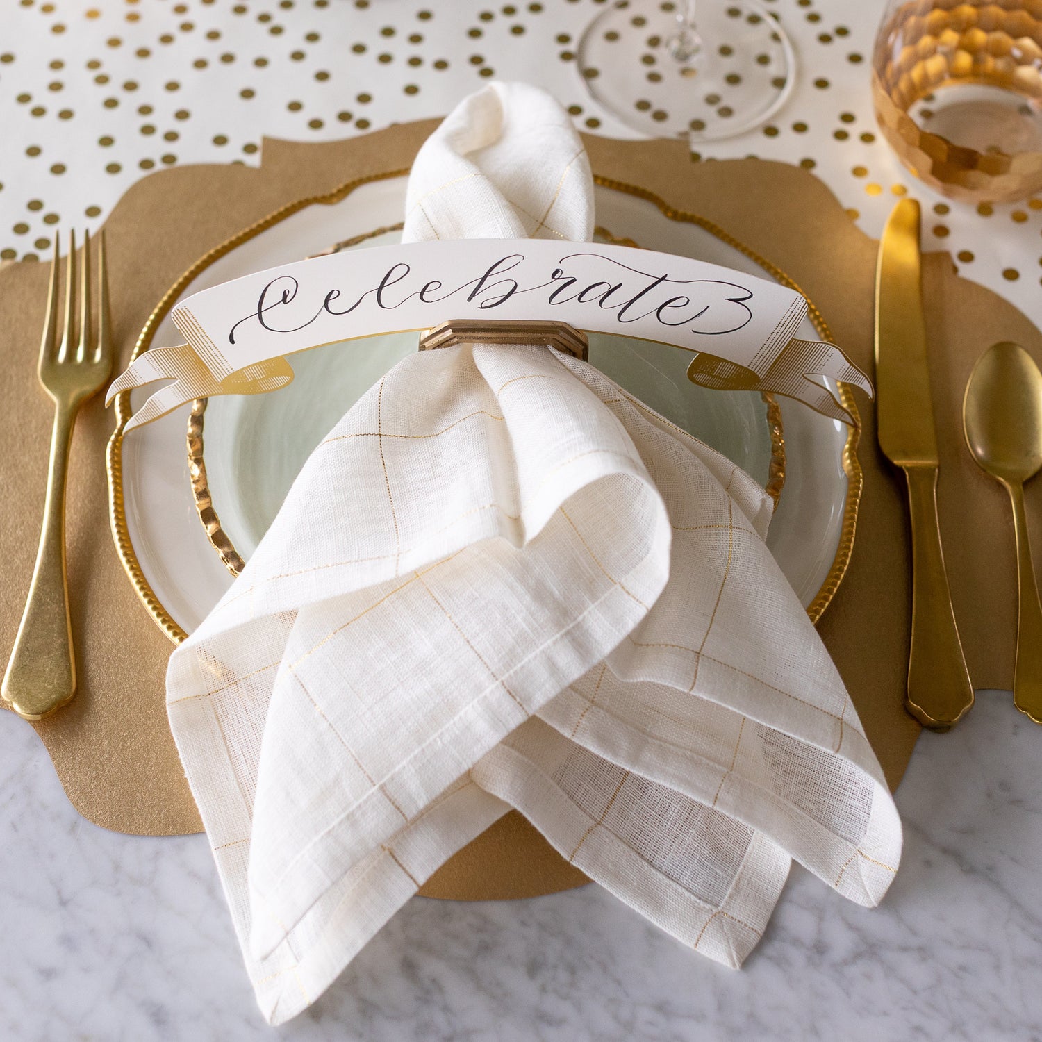 A place setting featuring the Brass Napkin Ring with Place Card Holder with a gold and white napkin in it, and Classic Gold Banner Table Accent reading "Celebrate" in the place card slot.