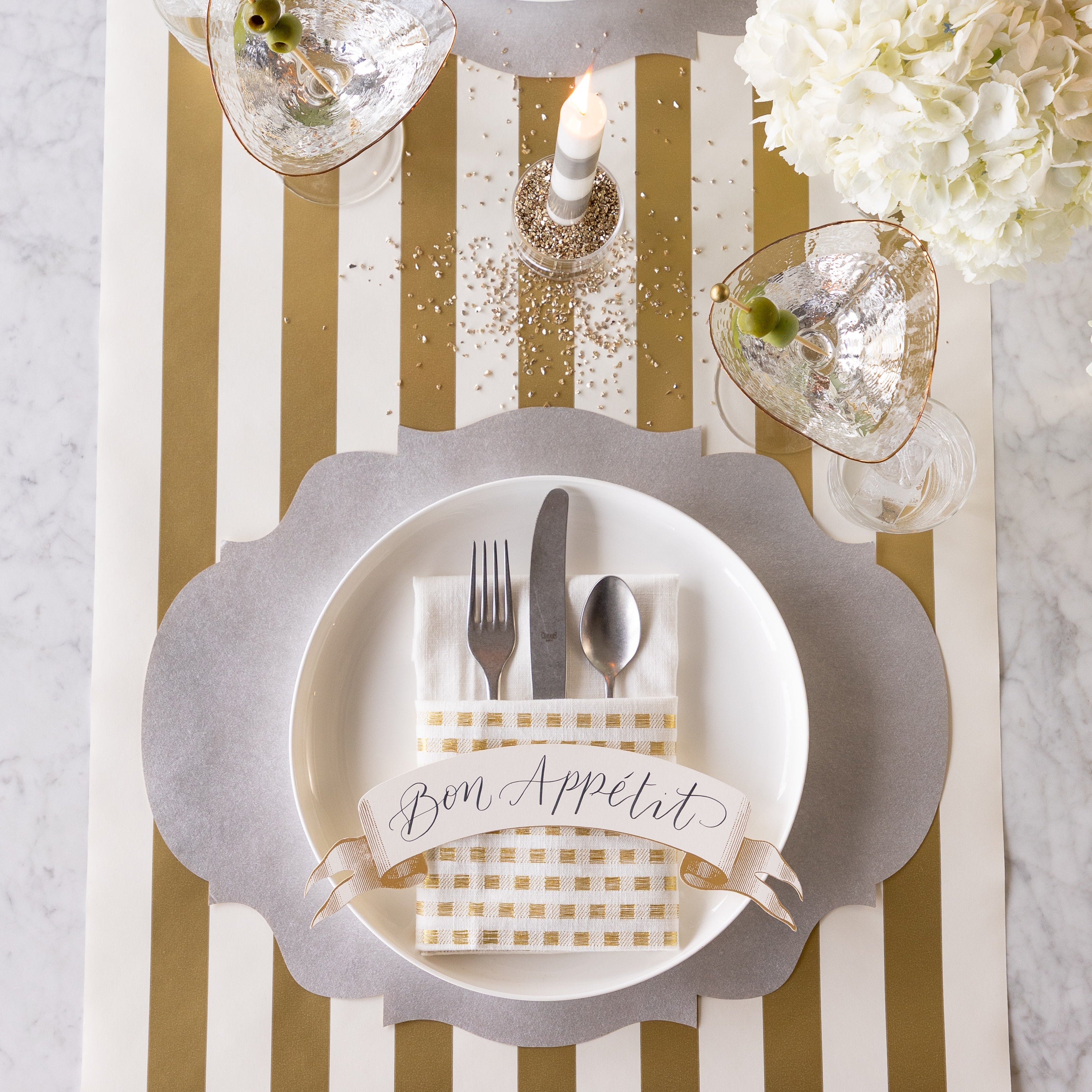 A place setting featuring the Die-cut Silver French Frame Placemat with silverware inside of a folded napkin and Classic Gold Banner Table Accent reading "Bon Appetit" on a dinner plate, a martini glass with olives, a lit candle, flowers and the Gold Classic Stripe Runner underneath.