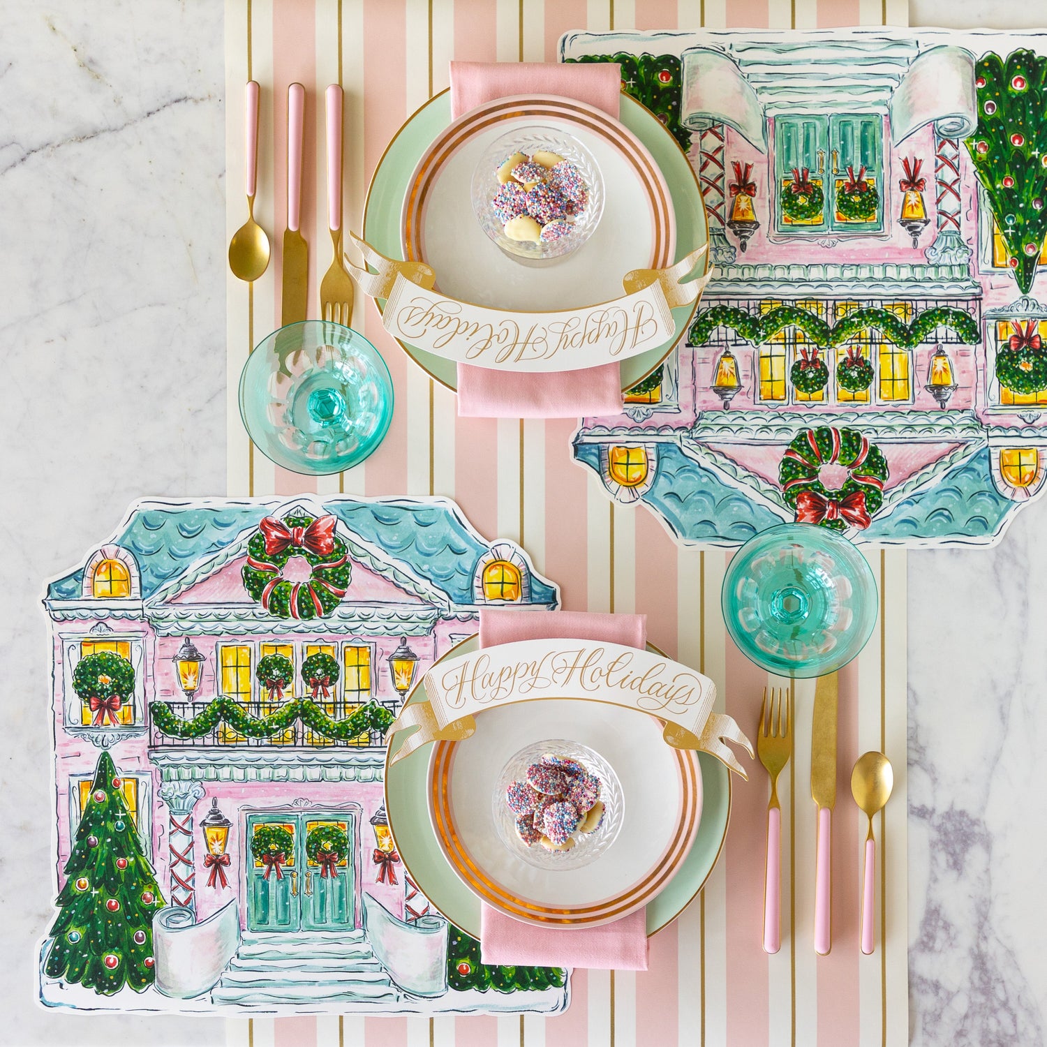 A table set with the Die-cut Holiday Home Placemats, seafoam and gold-rimmed plates, pink napkins, bowl of white chocolate buttons and Happy Holidays Banner Table Accent atop, and pink flatware on the Pink &amp; Gold Awning Stripe Runner.