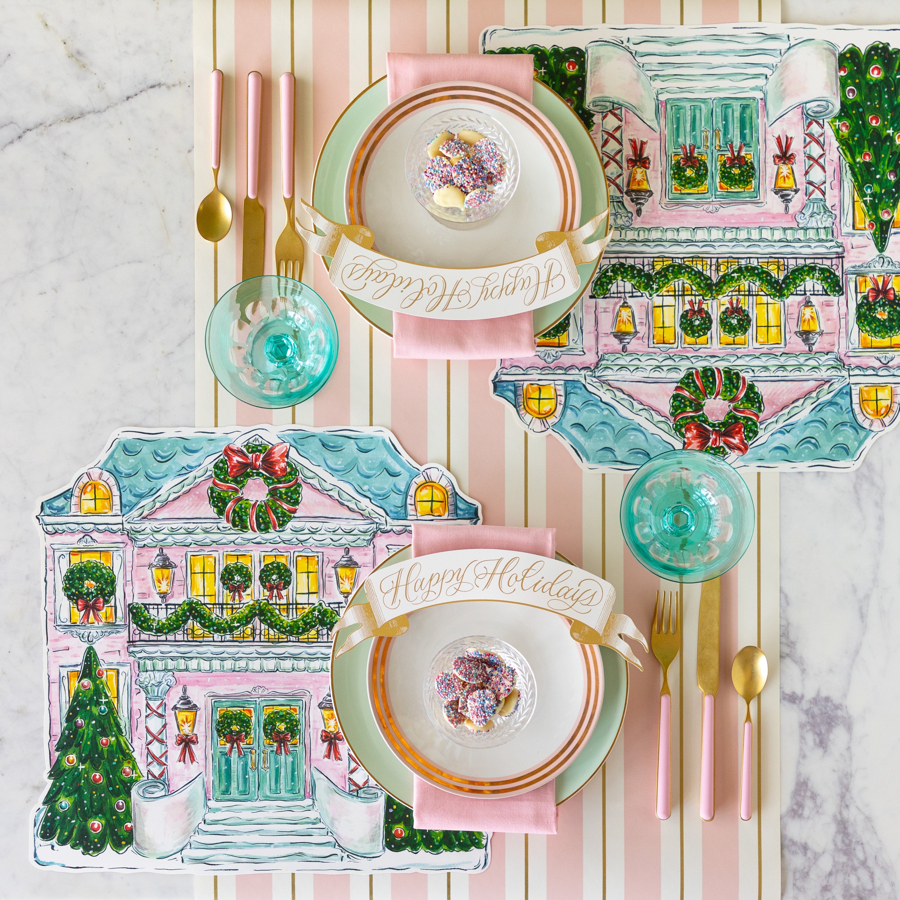 A table set with the Die-cut Holiday Home Placemats, seafoam and gold-rimmed plates, pink napkins, bowl of white chocolate buttons and Happy Holidays Banner Table Accent atop, and pink flatware on the Pink &amp; Gold Awning Stripe Runner.