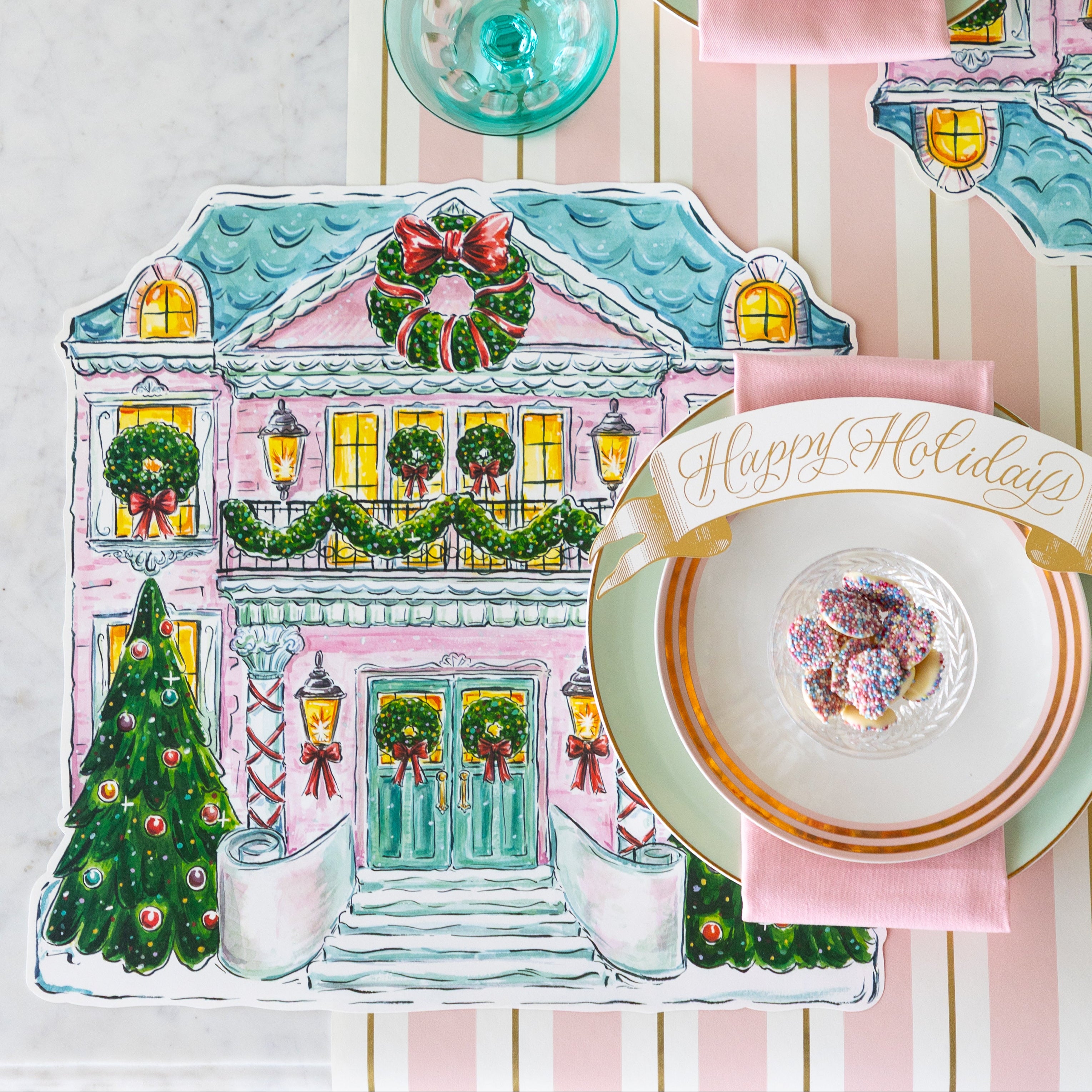 A place setting featuring the Die-cut Holiday Home Placemat, seafoam and gold-rimmed plates, a pink napkin, bowl of white chocolate buttons and Happy Holidays Banner Table Accent atop, on the Pink & Gold Awning Stripe Runner.