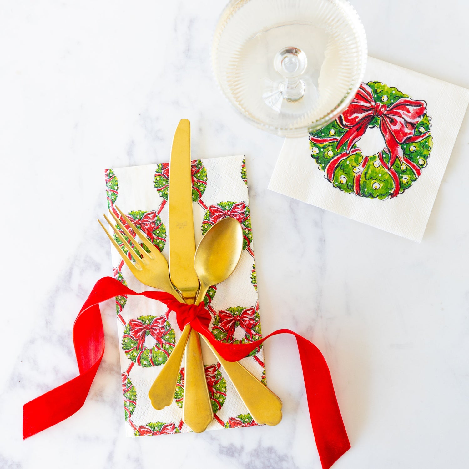 Holiday Wreath Guest Napkin with gold flatware tied together with a red ribbon on top, and a coupe glass on the Holiday Wreath Cocktail Napkin.