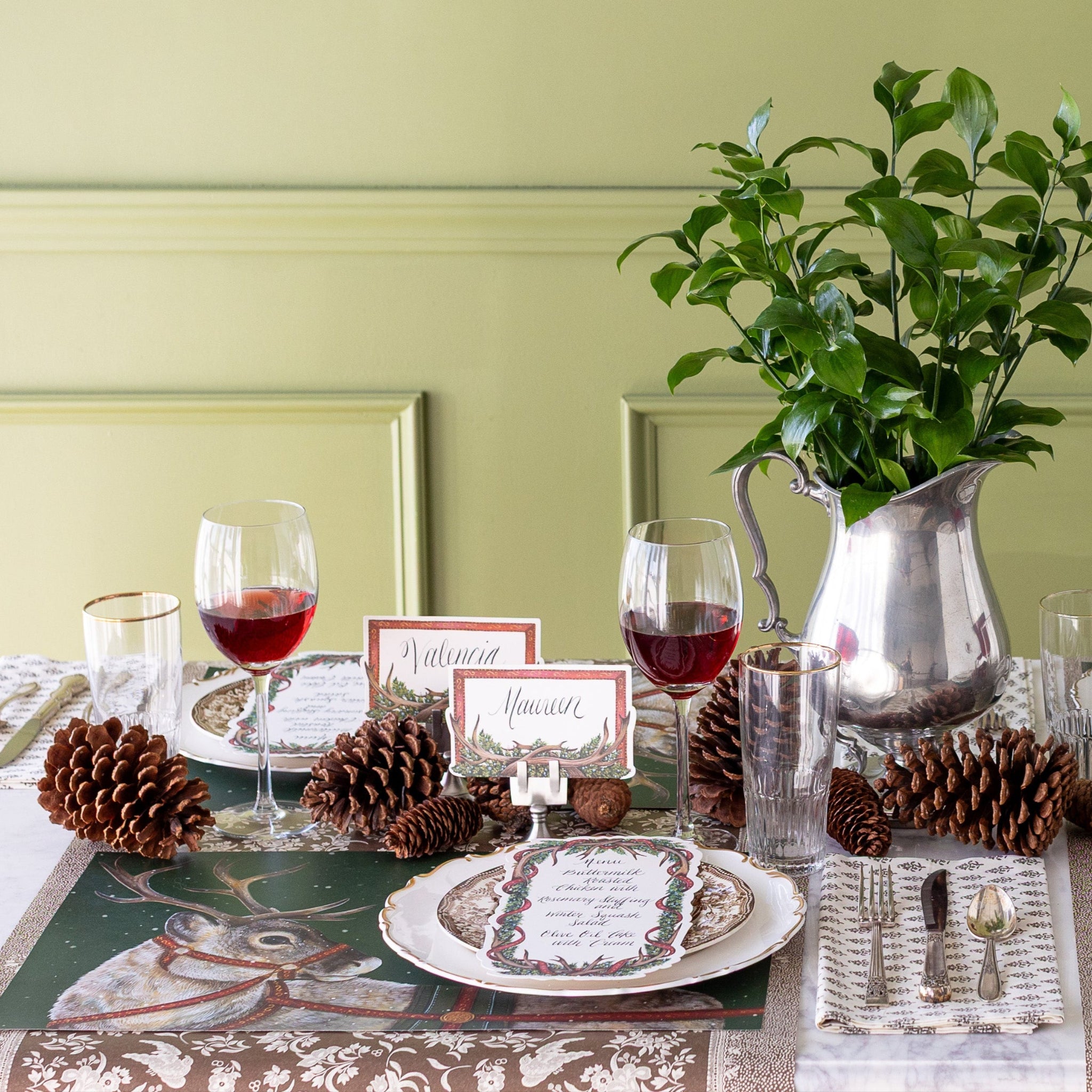 Decorative table setting with a floral centerpiece, pinecones, wine glasses, Holiday Reindeer Placemats, Antler Garland Table Cards, and Antler Frame Place Cards on the Brown Regal Peacock Runner.