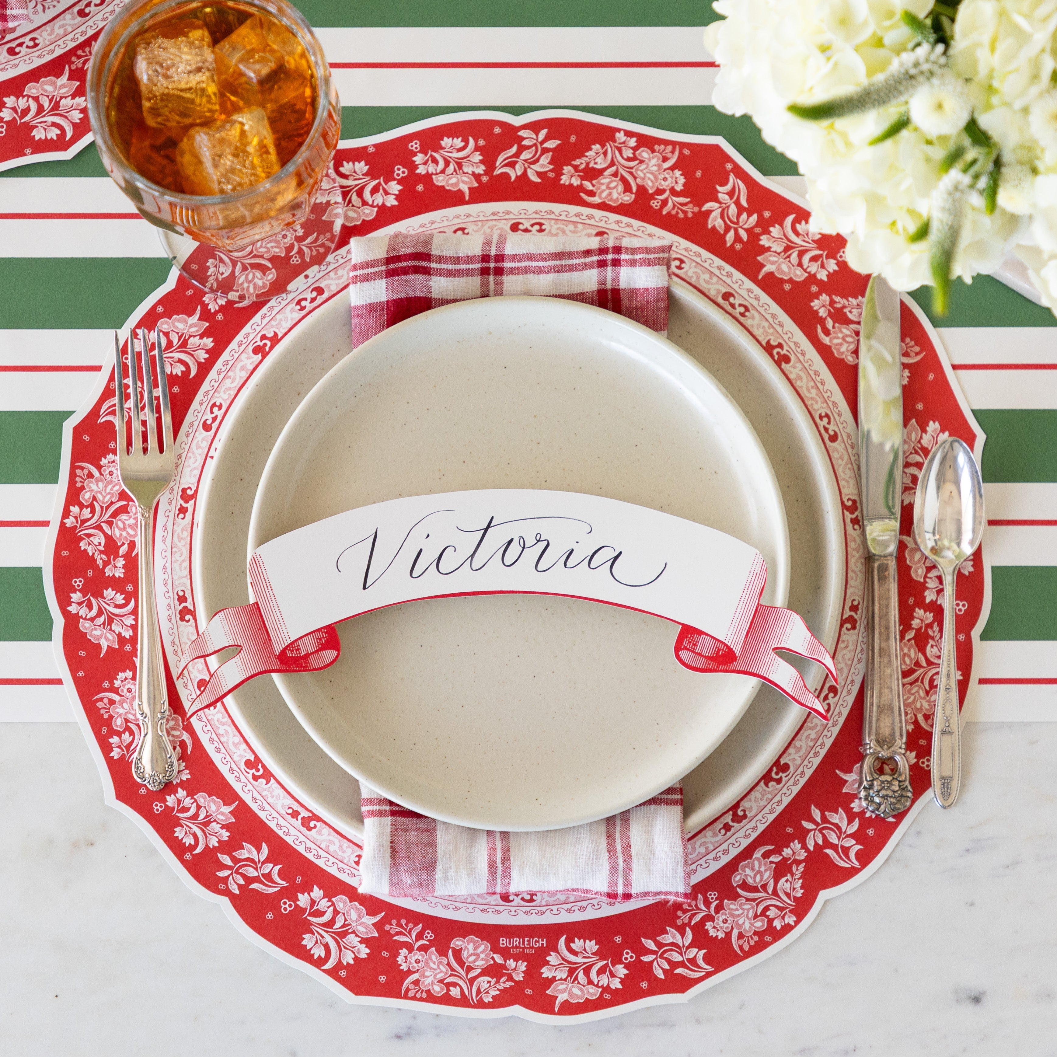 A place setting featuring the Die-cut Red Regal Peacock Placemat, off-white dinner plates with a red-and-white checkered napkin in between and Classic Red Banner Table Accent reading "Victoria" atop, vintage flatware, a glass of tea, and Green & Red Awning Stripe Runner underneath.