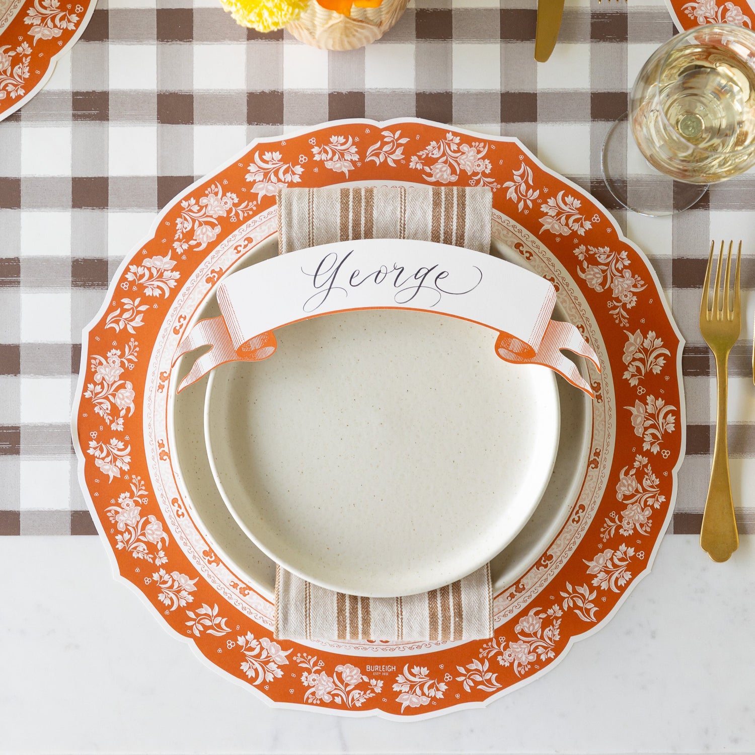 An elegant place setting featuring the Die-cut Pumpkin Regal Placemat, off-white plates, brown-and-cream striped napkin with the Classic Orange Banner Table Accent atop, on the Brown Painted Check Runner.