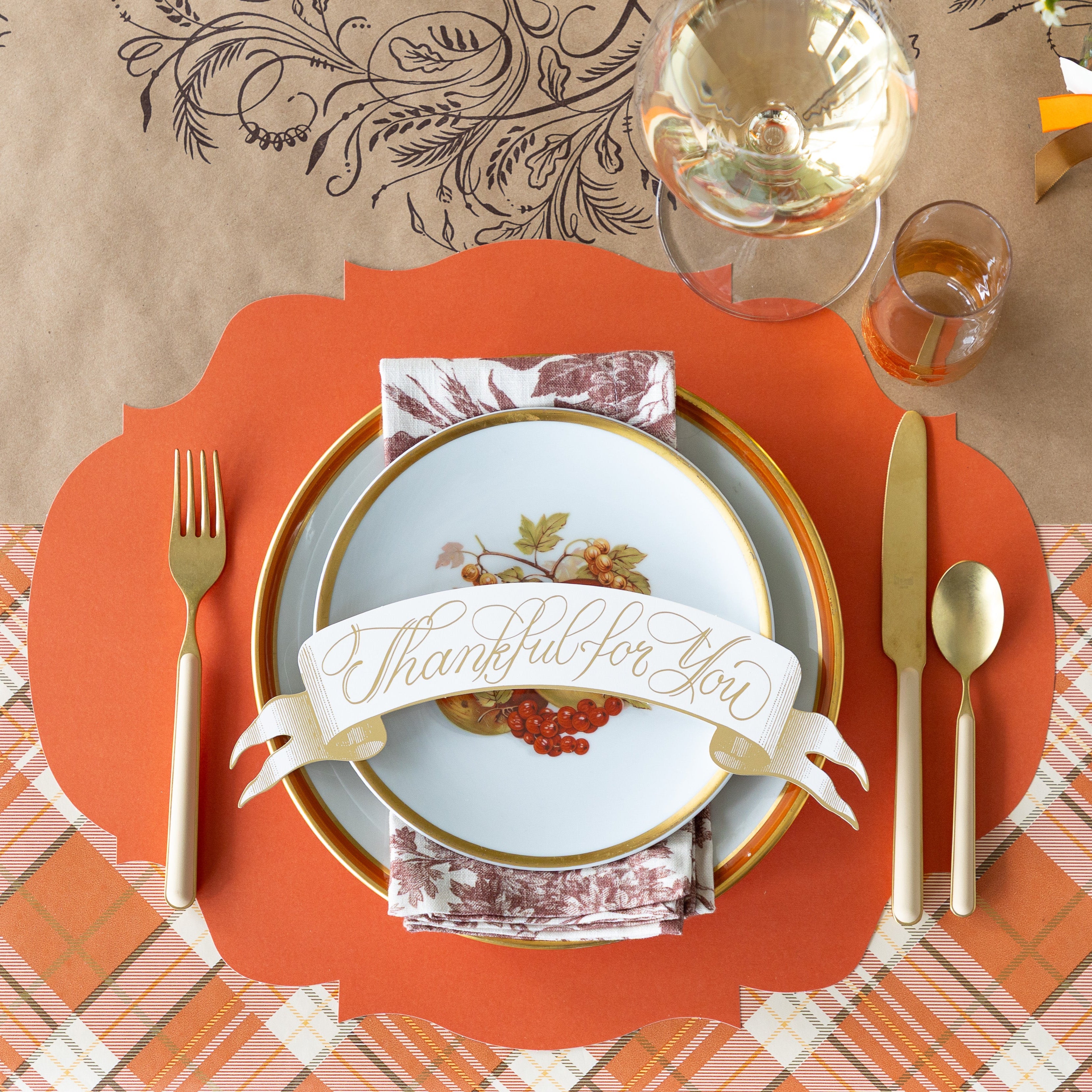 A fall table setting featuring the Orange French Frame Placemat with vintage plates, a floral napkin and Thankful for you Banner Table Accent atop, tan flatware, and a wine glass on the Kraft Thankful Runner.
