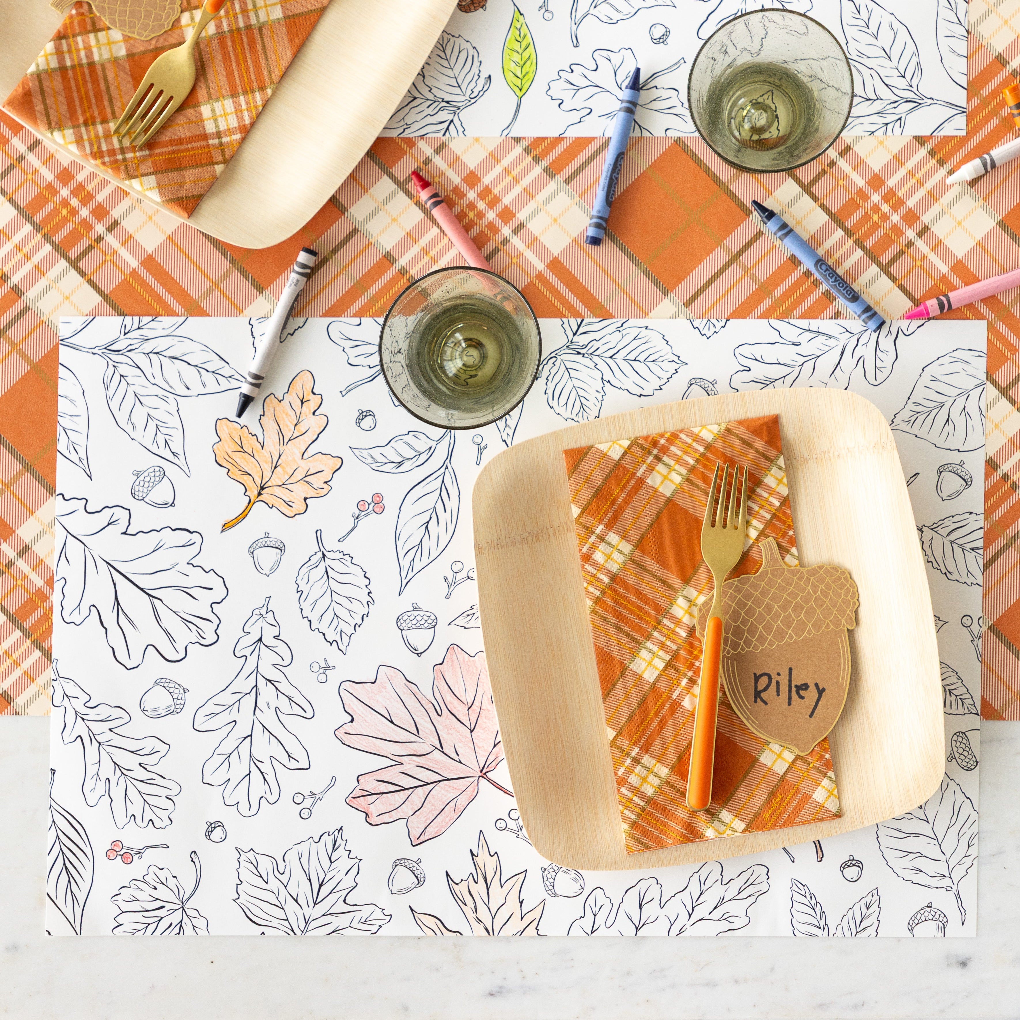 A place setting featuring the Coloring Leaves Placemat partially colored in, a paper plate with the Orange Plaid Guest Napkin, orange fork and Acorn Place Card reading "Riley" on top and scattered crayons on the Orange Plaid Runner.