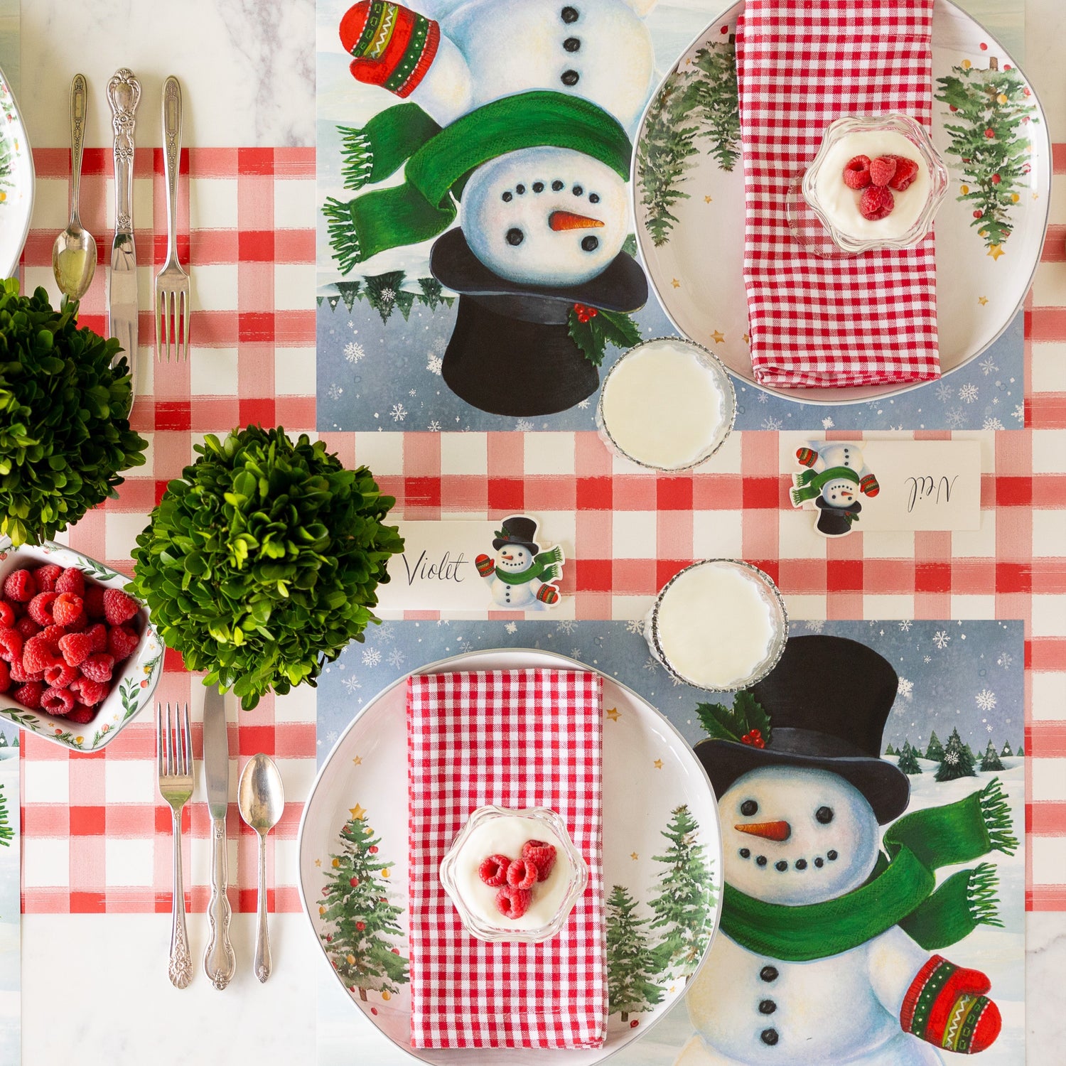 Two place settings with Jolly Snowman Placemats, red checkered napkins, parfait desserts topped with raspberries, and glasses of milk arranged on the Red Painted Check Runner.