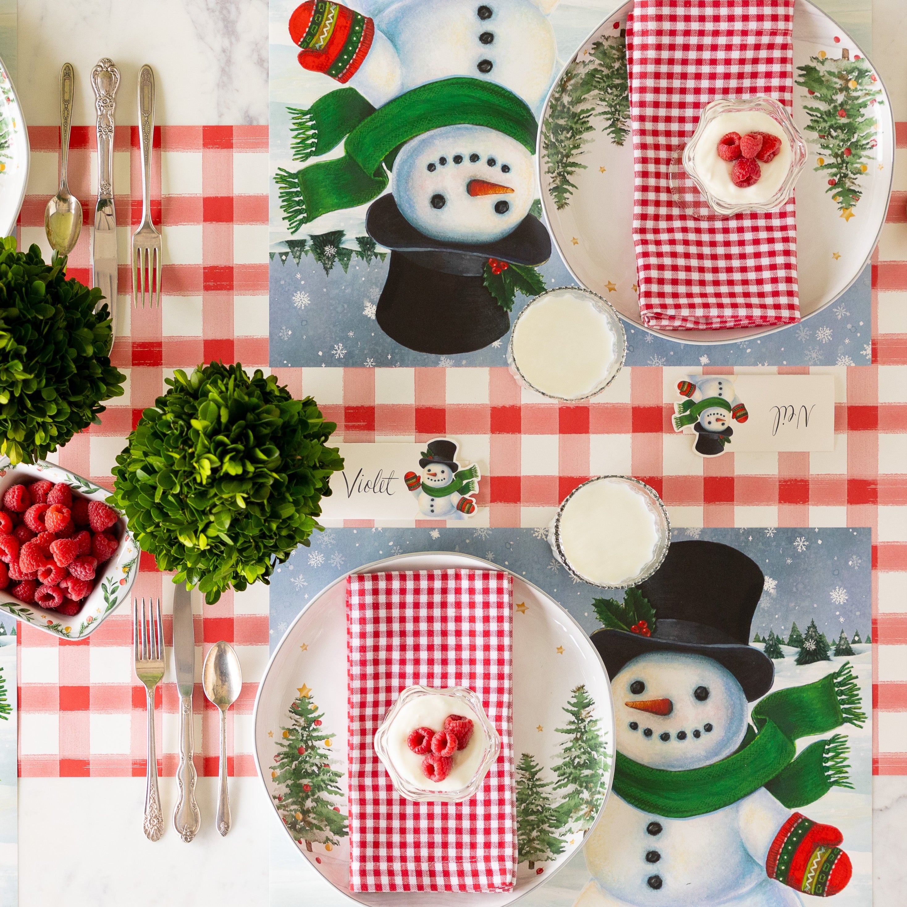 Two place settings with Jolly Snowman Placemats, red checkered napkins, parfait desserts topped with raspberries, and glasses of milk arranged on the Red Painted Check Runner.