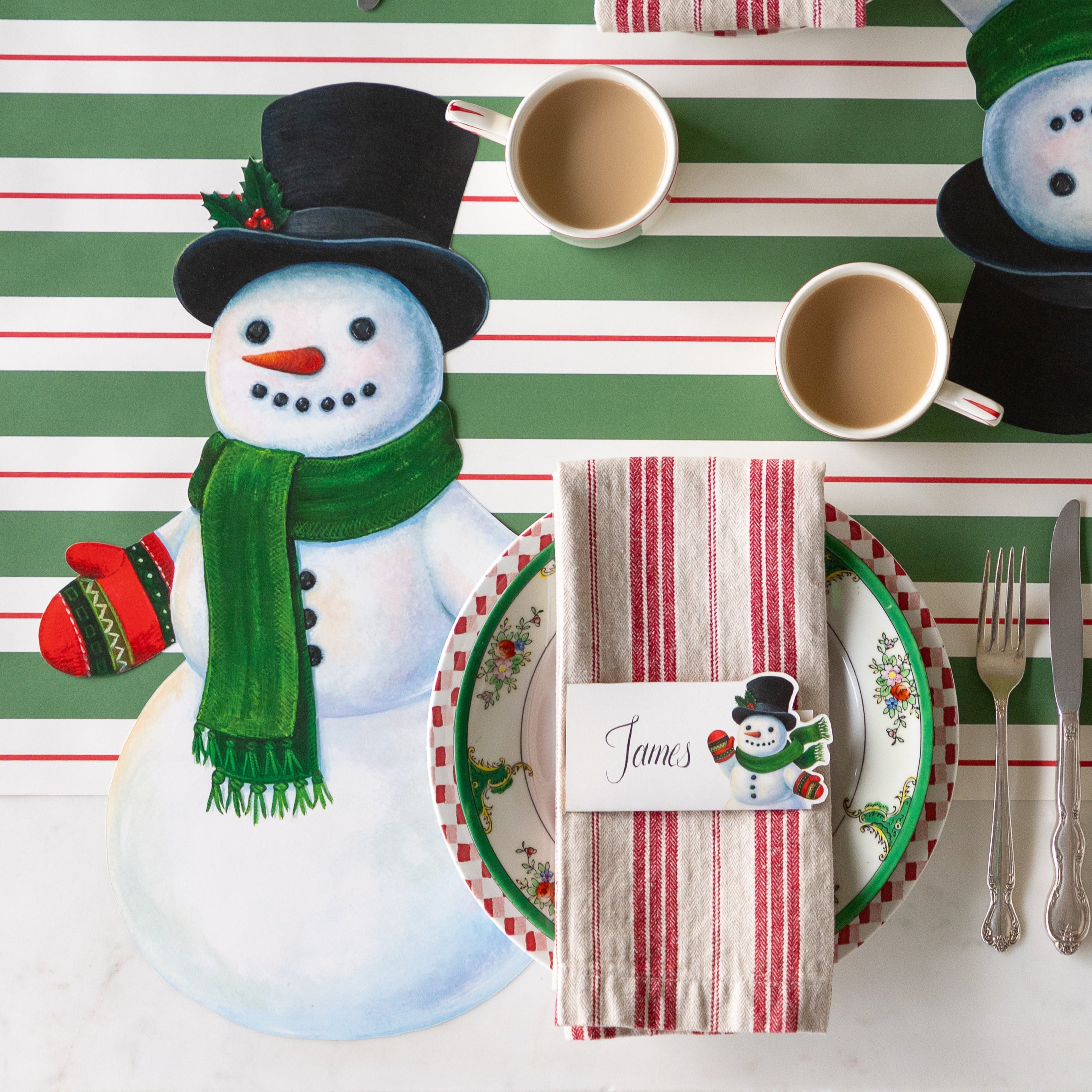 A festive place setting with the Die-cut Jolly Snowman Placemat, cup of coffee, red striped napkins with the Jolly Snowman Place Card on top and Green & Red Awning Stripe Runner underneath.