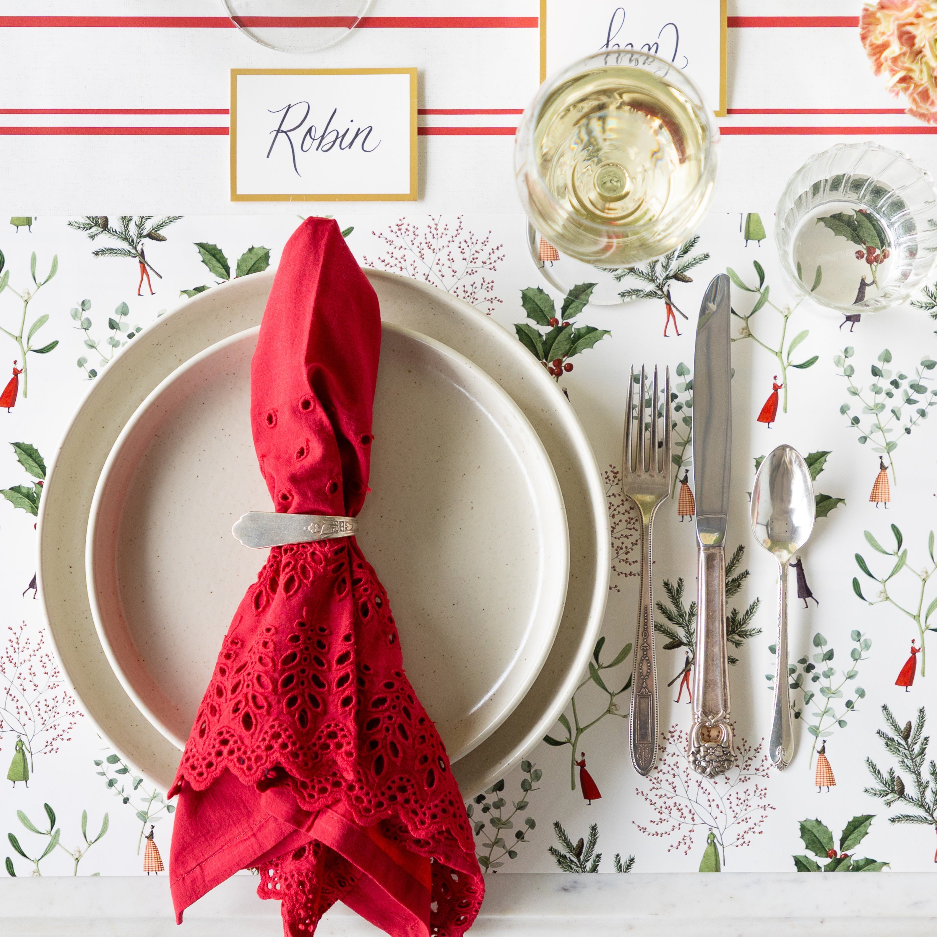 A place setting featuring the Happy Christmas Placemat, dinner plates with a red lace napkin in a holder atop, vintage silverware, a glass of wine and water, Gold Frame Place Card reading "Robin", and Antique Red Stripe Runner underneath.
