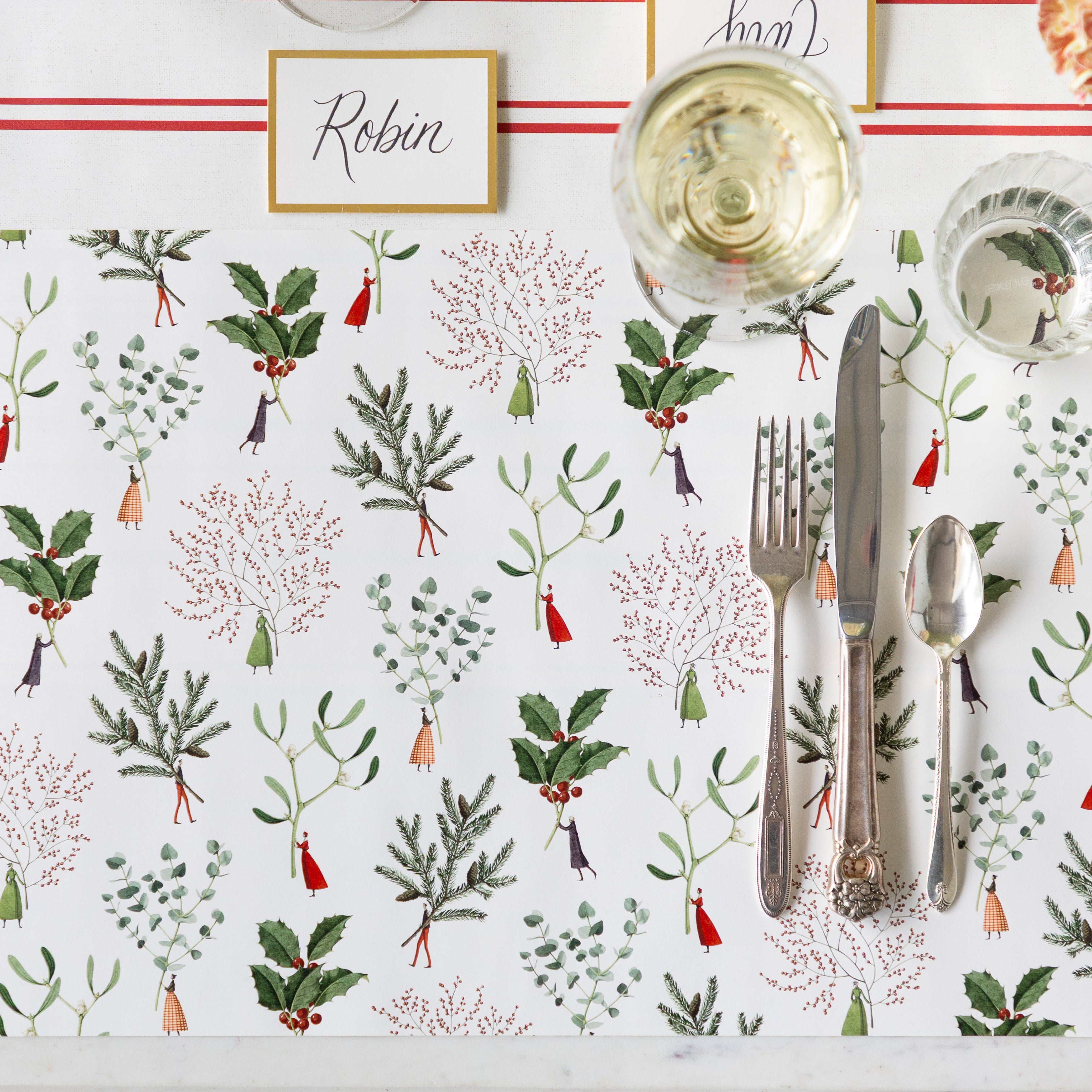A place setting featuring the Happy Christmas Placemat with vintage silverware, a glass of wine and water atop, Gold Frame Place Card reading "Robin" behind, and Antique Red Stripe Runner underneath.