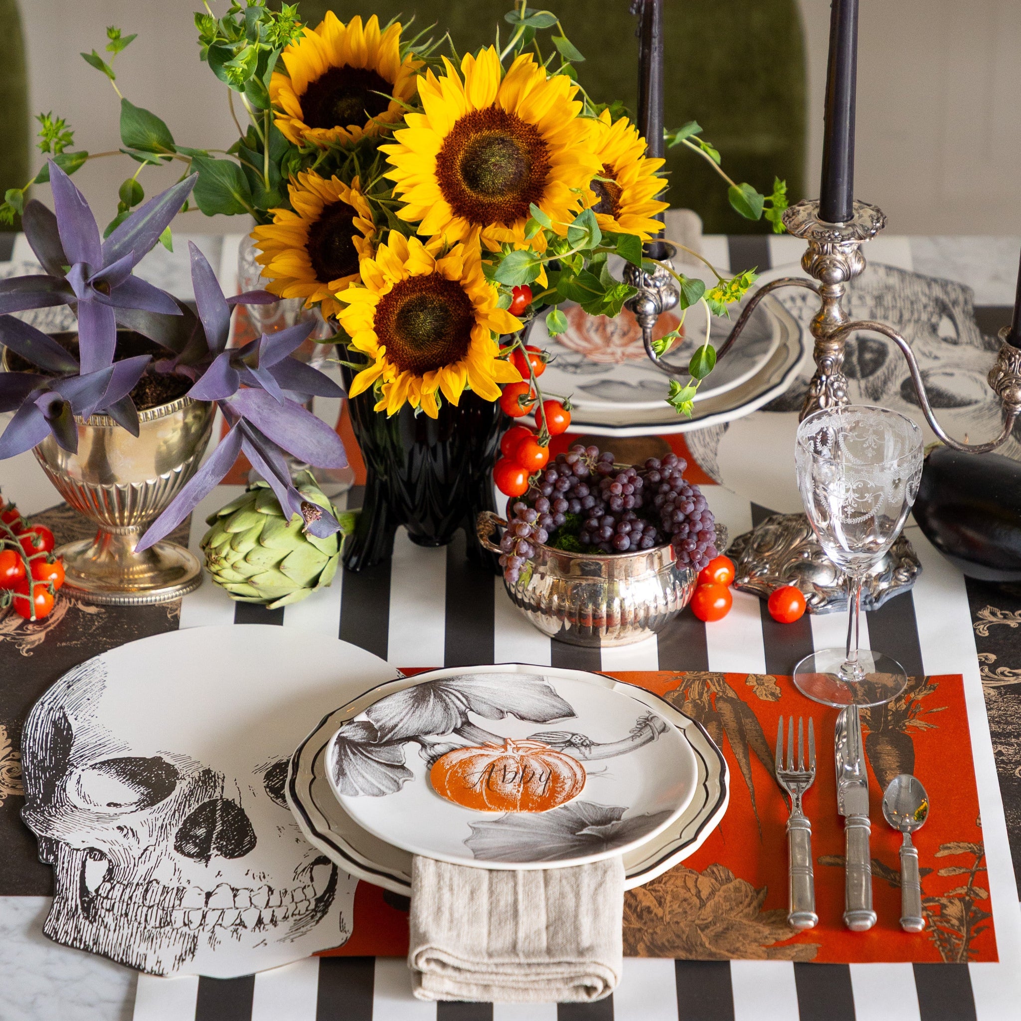 Halloween-themed table setting with the Die-cut Skull Placemat, Pumpkin Place Card,Harvest Vegetable Persimmon Placemat, sunflowers and fruits &amp; veggies on the Black Classic Stripe and Archival Black Runner.
