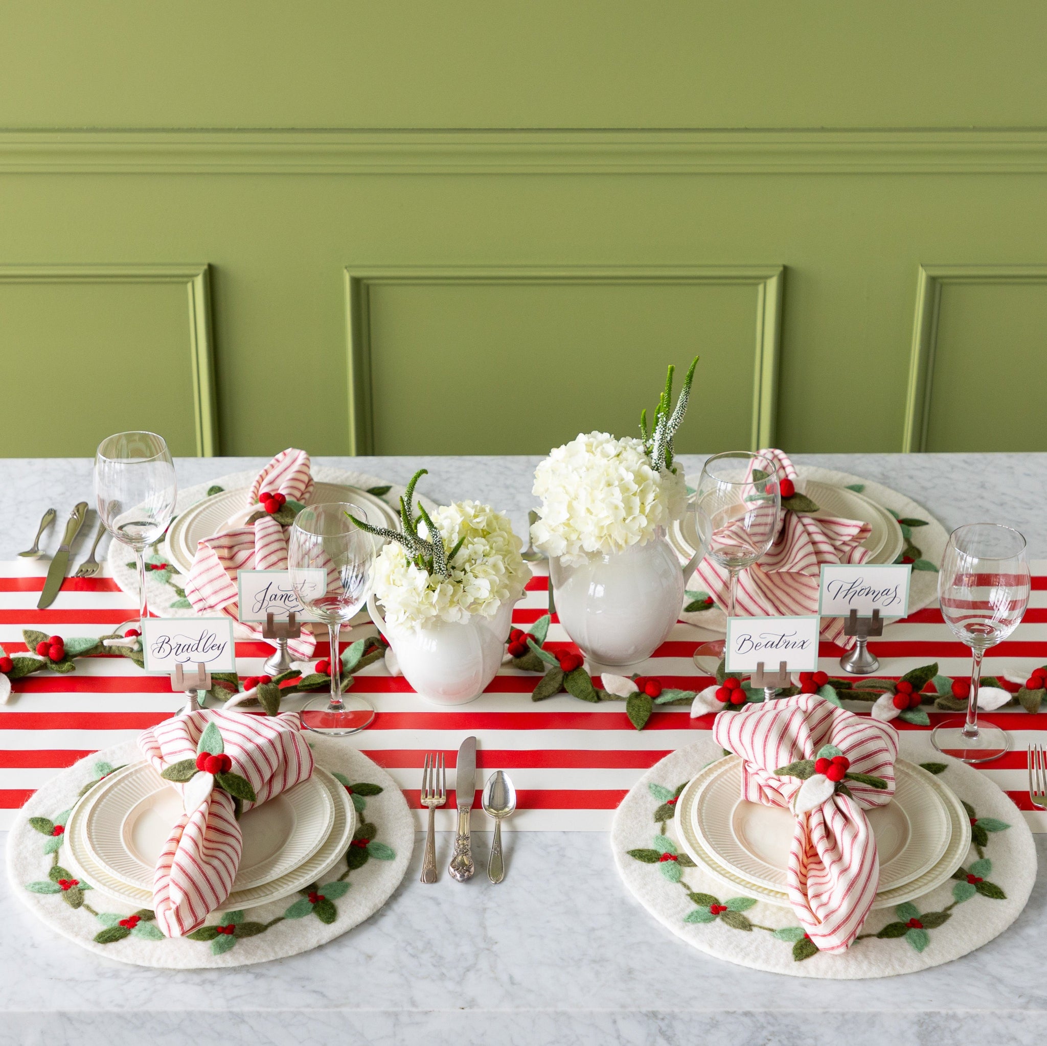 A festive table setting featuring the Felt Berry Garland in the center, flowers, Berry Wreath Placemat, white dinnerware, red-and-white striped napkins in the Felt Berry Napkin Ring and Seafoam Frame Place Cards on the Red Classic Stripe Runner.