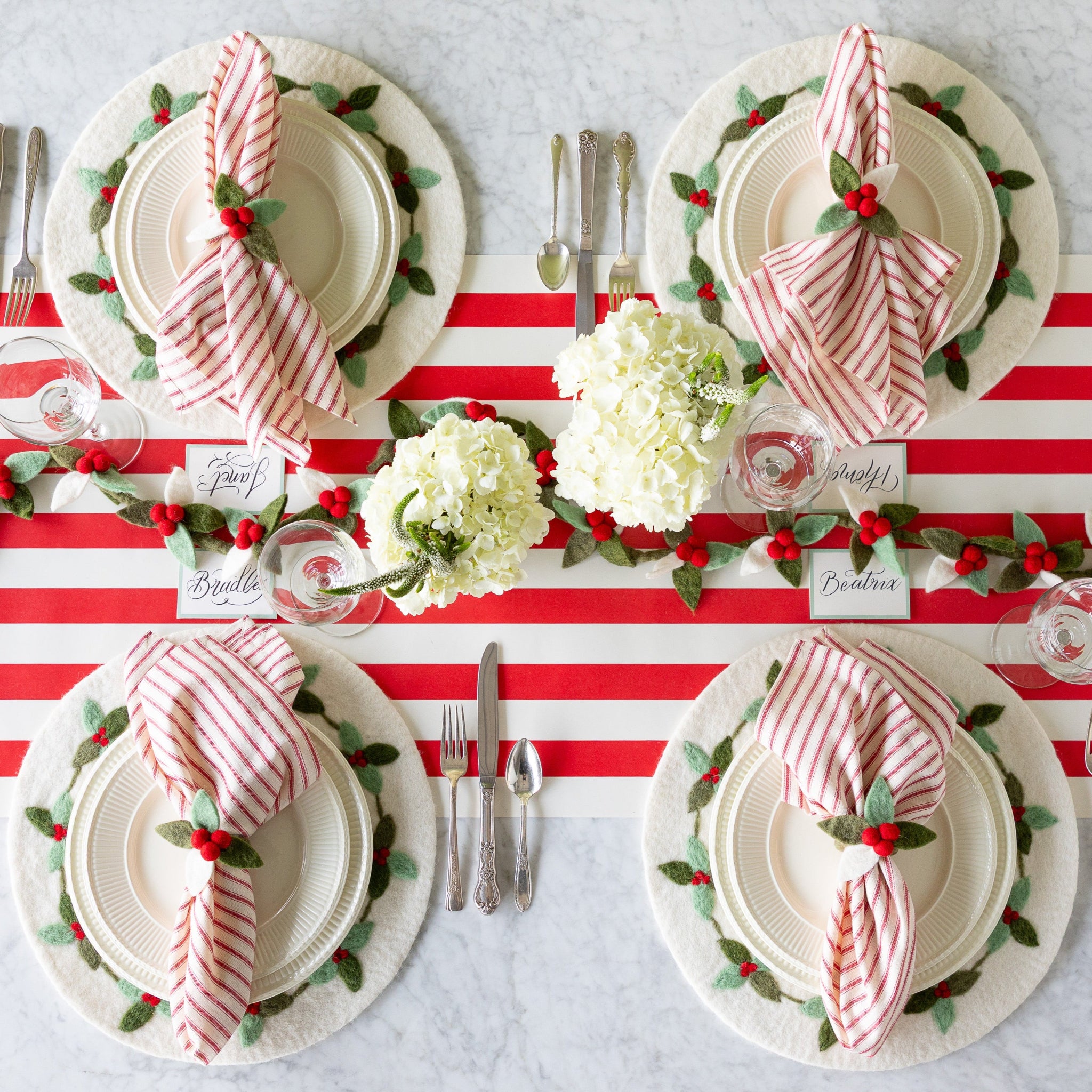 A festive table setting featuring the Felt Berry Garland in the center, flowers, Berry Wreath Placemat, white dinnerware, red-and-white striped napkins in the Felt Berry Napkin Ring and Seafoam Frame Place Cards on the Red Classic Stripe Runner.