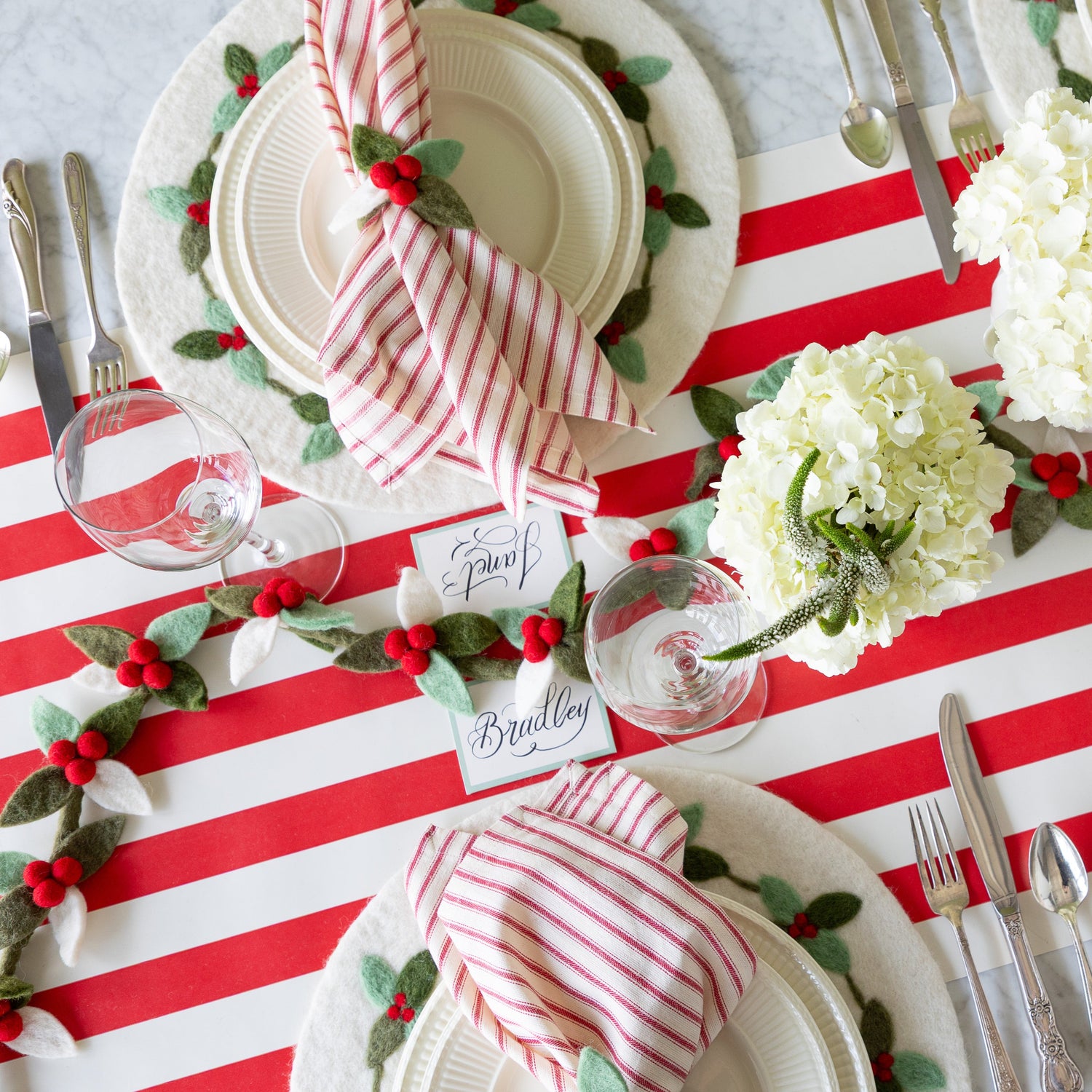 A festive table setting featuring the Felt Berry Garland in the center, flowers, Berry Wreath Placemat, white dinnerware, red-and-white striped napkins in the Felt Berry Napkin Ring and Seafoam Frame Place Cards on the Red Classic Stripe Runner.