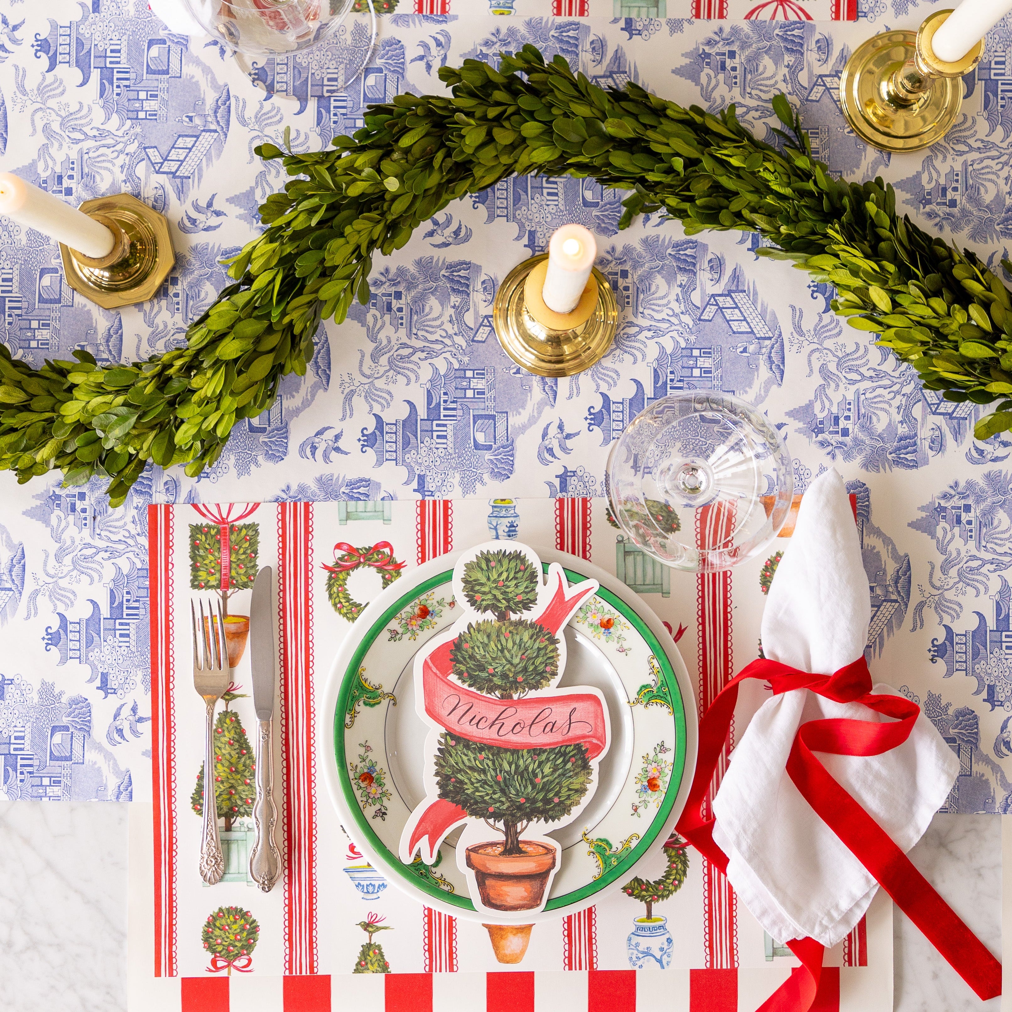 An elegant table setting with a topiary garland featuring the Holiday Topiary Placemat, a red ribbon tied around a napkin and Red Classic Stripe Runner running vertically and Blue Regal Peacock Runner running horizontally underneath.