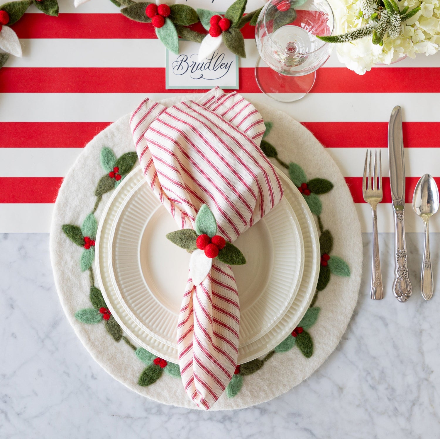 A festive place setting featuring the Felt Mistletoe Berry Placemat with white dinner plates and a red-and-white striped napkin in the Felt Berry Napkin Ring atop, vintage silverware, a wine glass, and Seafoam Frame Place Card reading "Bradley", on the Red Classic Stripe Runner.