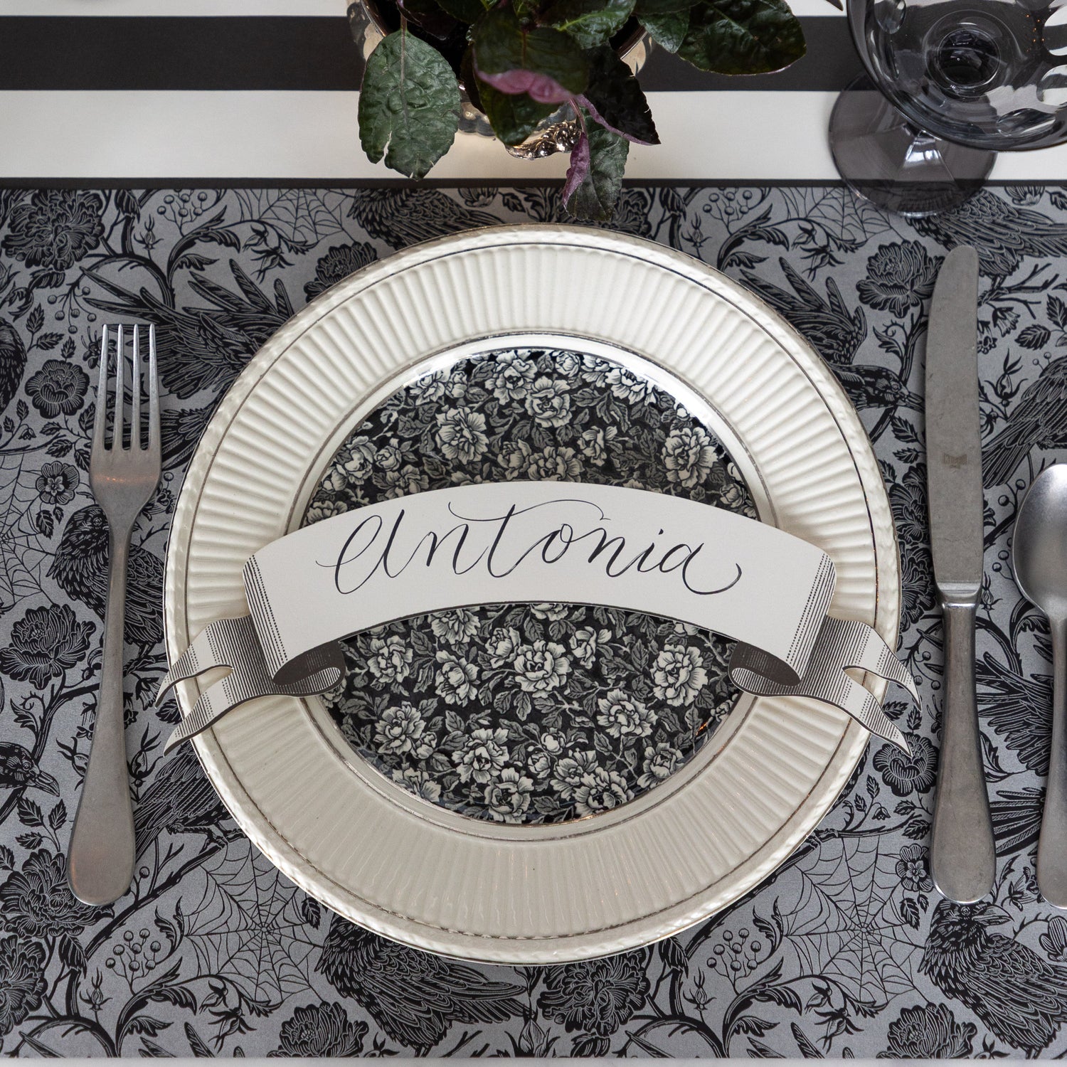 A spooky place setting featuring the Raven Toile Placemat with silver flatware, a dinner and salad plate with the Classic Black Banner Table Accent with &quot;Antonia&quot; written on it on top and Black Classic Stripe Runner underneath.