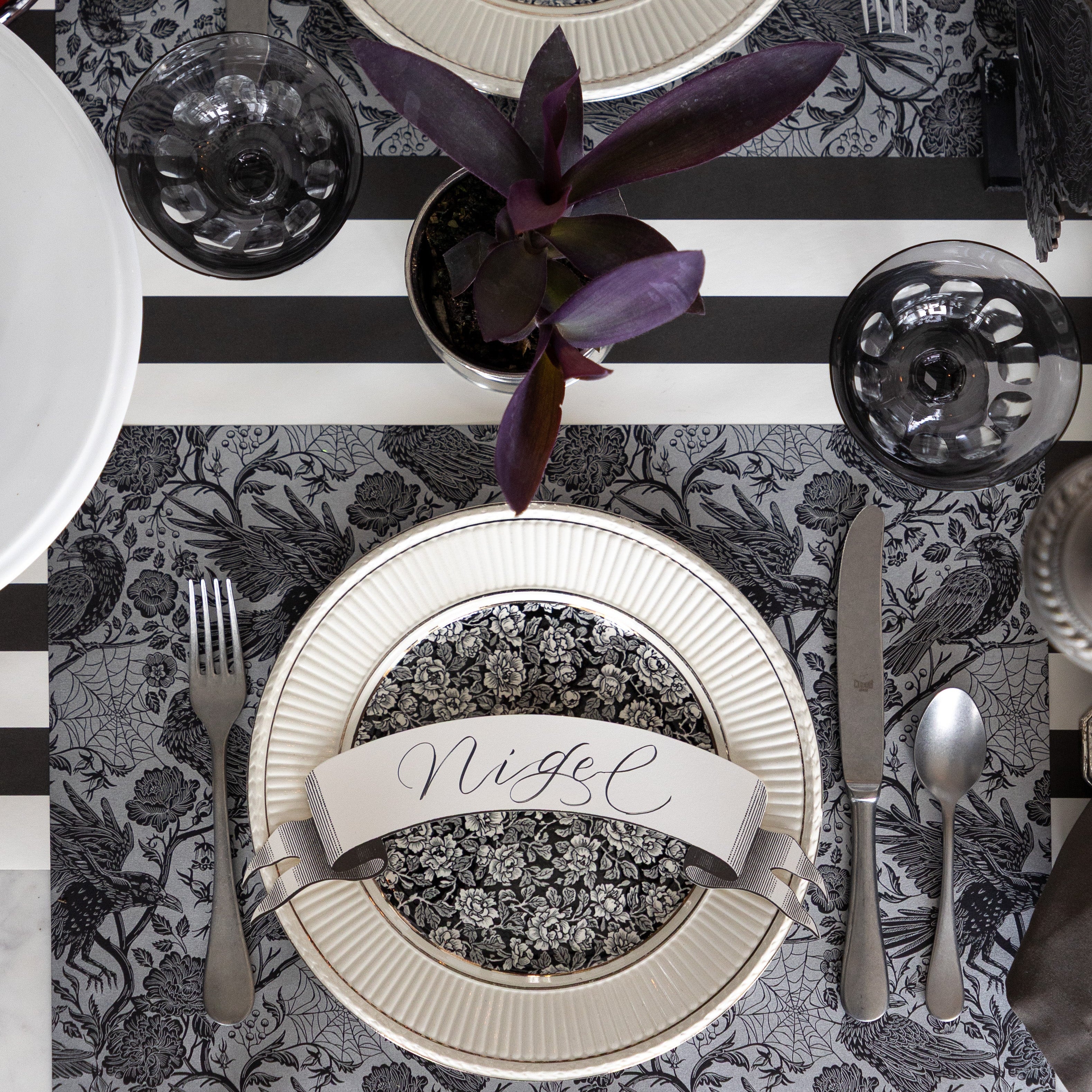 A spooky place setting featuring the Raven Toile Placemat with silver flatware, a dinner and salad plate with the Classic Black Banner Table Accent with "Nigel" written on it on top and Black Classic Stripe Runner underneath.