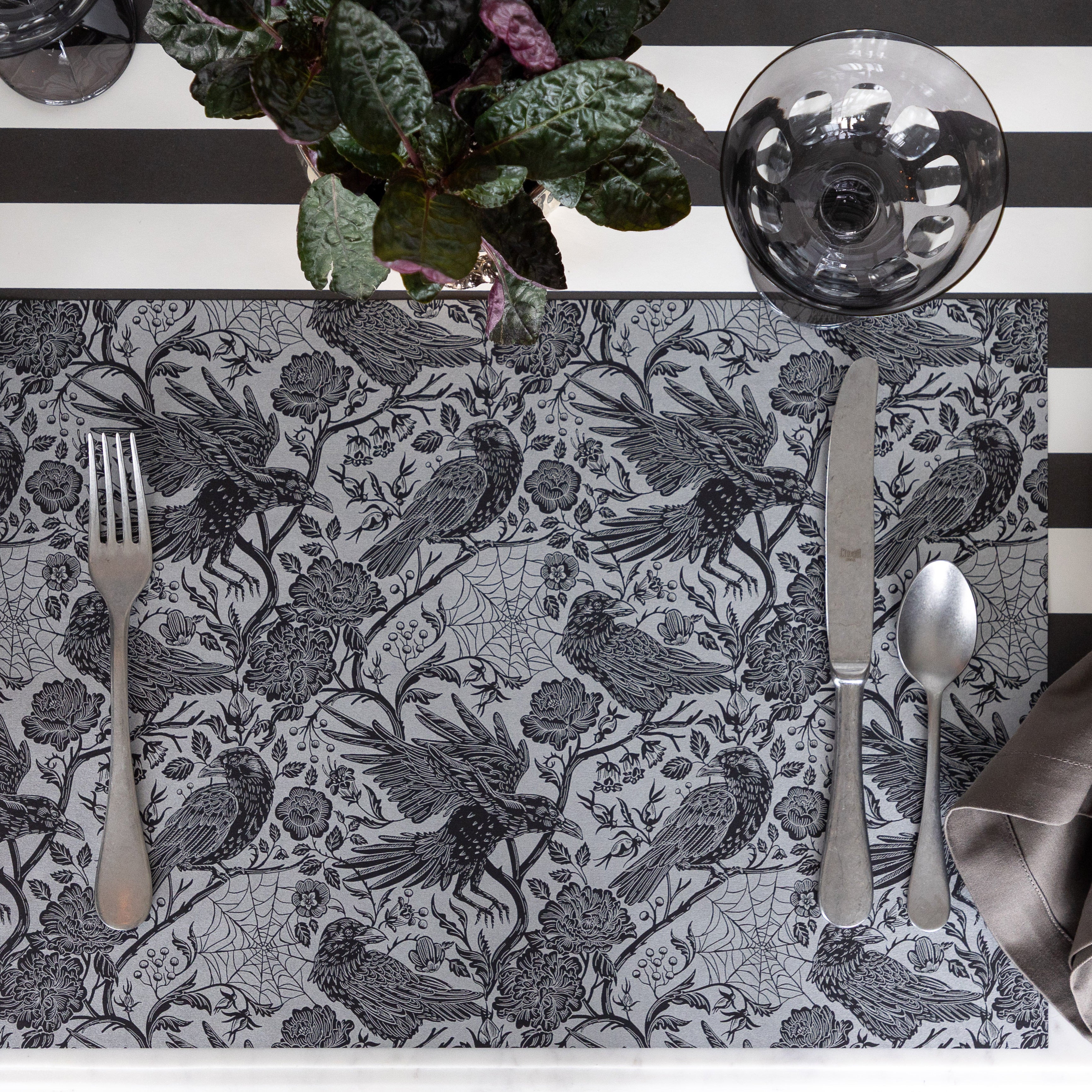 A place setting with only flatware to show the Raven Toile Placemat on the Black Classic Stripe Runner and Smoke Rialto Coupe glass and plant nearby.