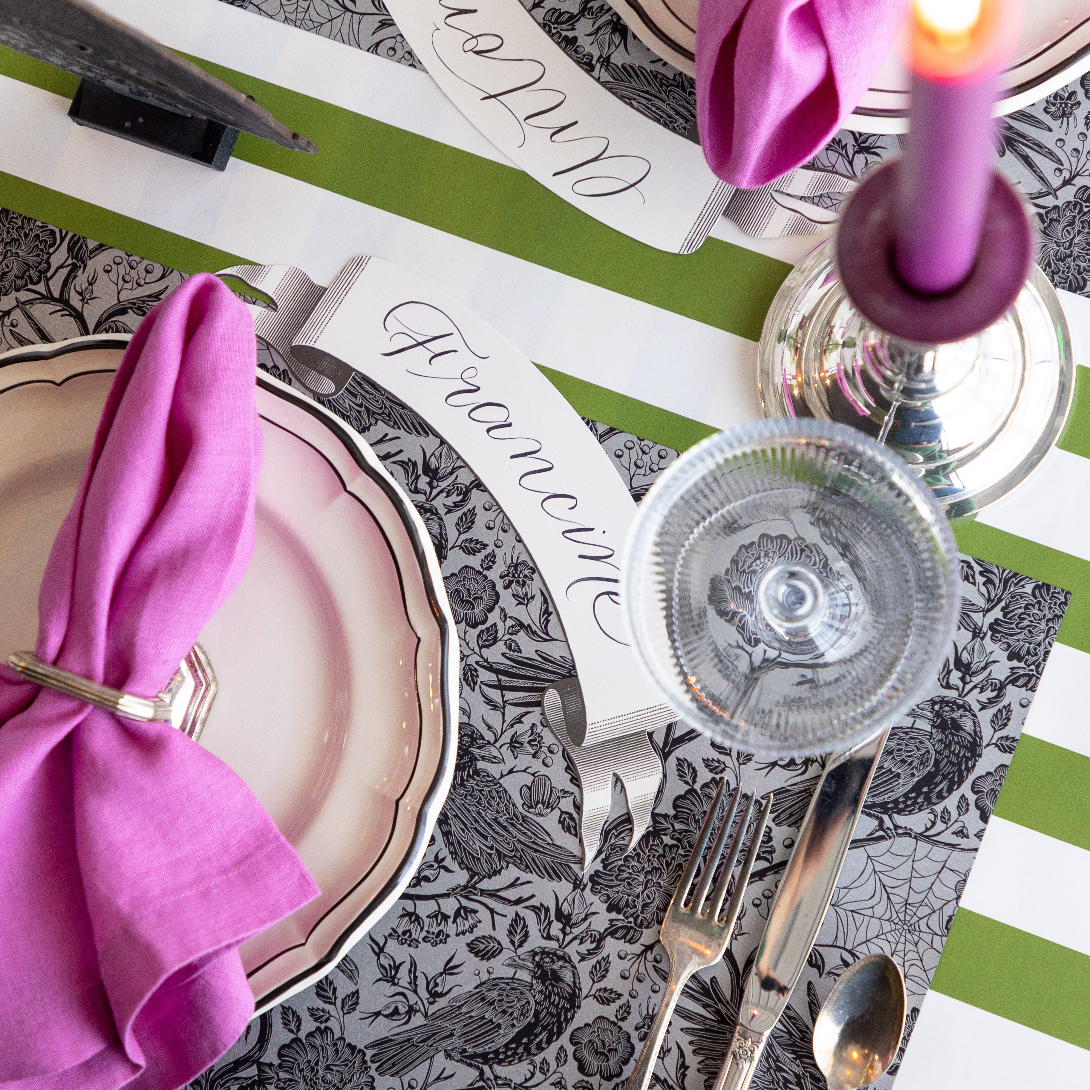 A place setting featuring the Raven Toile Placemat, Black Banner Table Accent reading "Francine" beside the plate, magenta napkin on the plate with a matching candle in a holder beside it and Moss Classic Stripe Runner underneath.
