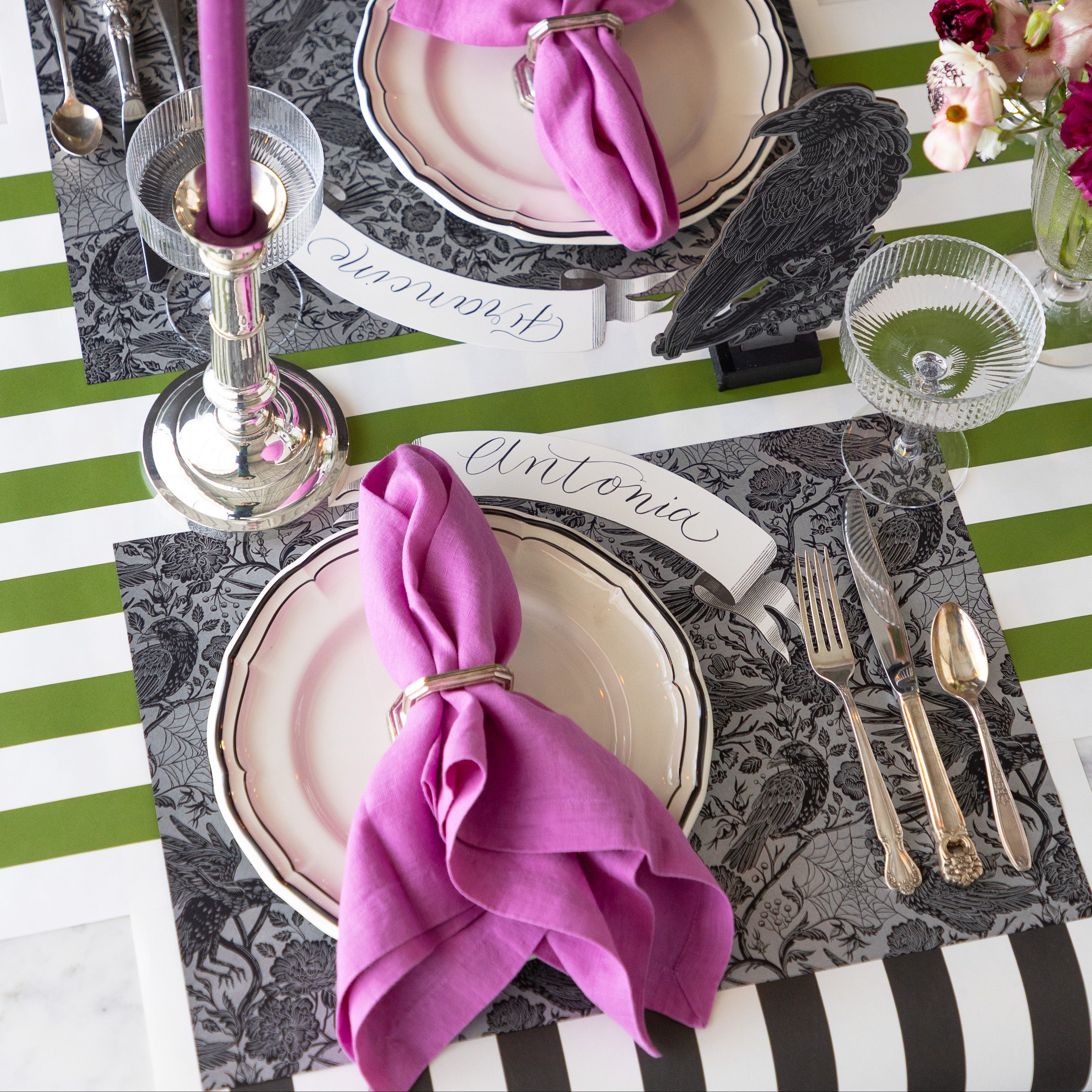 A place setting featuring the Raven Toile Placemat, Raven Ornament, Black Banner Table Accent reading "Antonia" beside the plate and Moss and Black Classic Stripe Runners underneath.