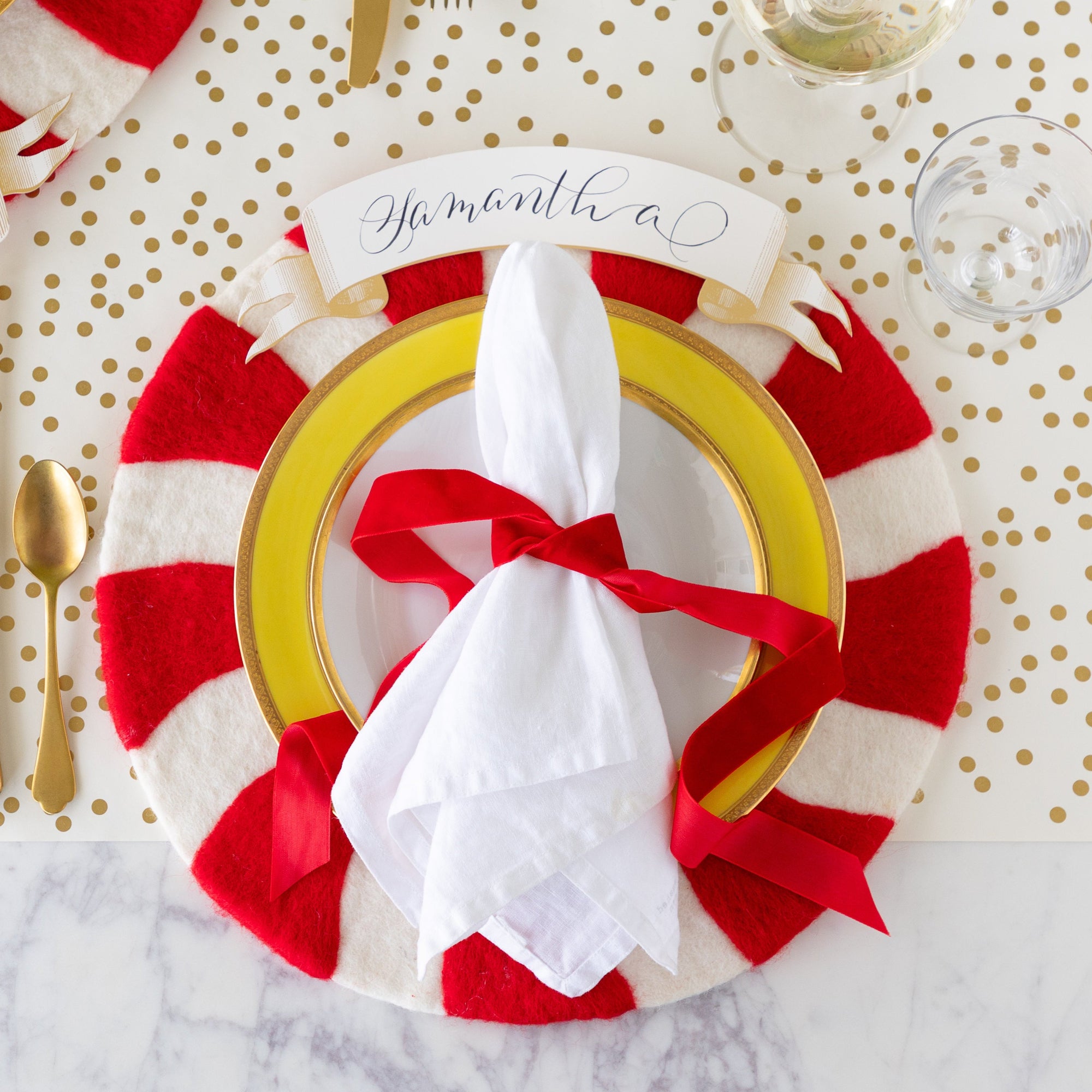 A place setting featuring the Red Felt Candy Stripe Placemat, yellow and gold rimmed plates with a white napkin tied in a red velvet ribbon atop, Classic Gold Banner Table Accent reading &quot;Samantha&quot; above, and Gold Confetti Runner underneath.