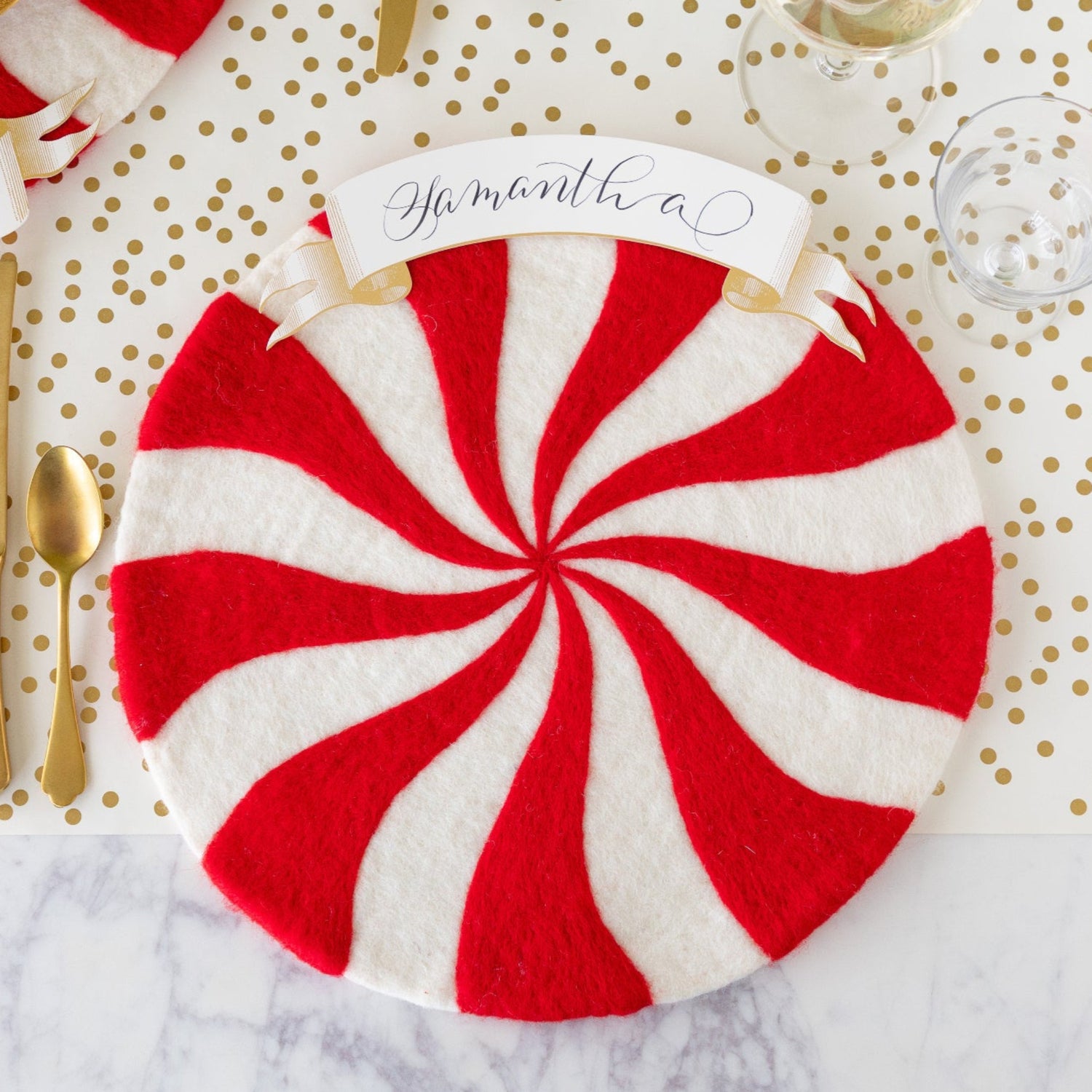 The Red Felt Candy Stripe Placemat with the Classic Gold Banner Table Accent reading "Samantha" atop, gold flatware beside it, an empty glass, and Gold Confetti Runner underneath.