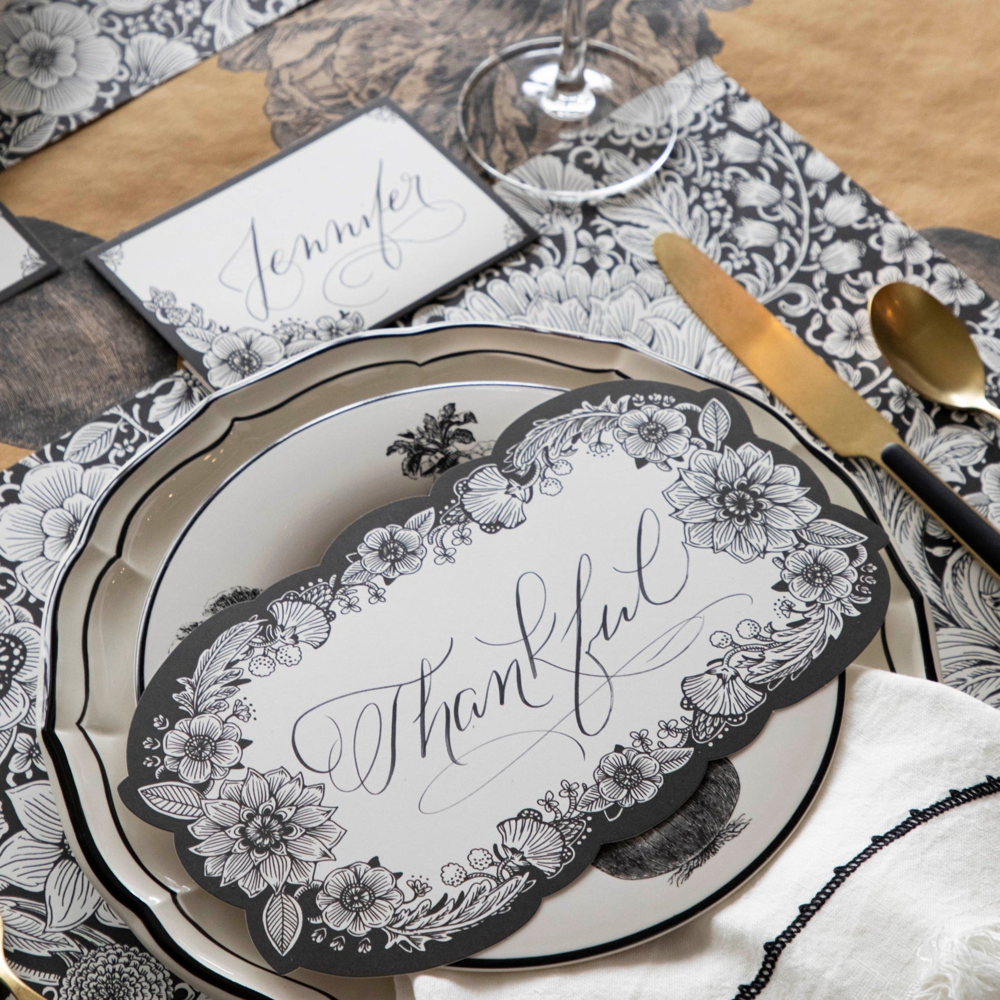 Close-up of an elegant place setting featuring an Ebony Harvest Blooms Table Card with "Thankful" written on it in lovely script resting on the plate.