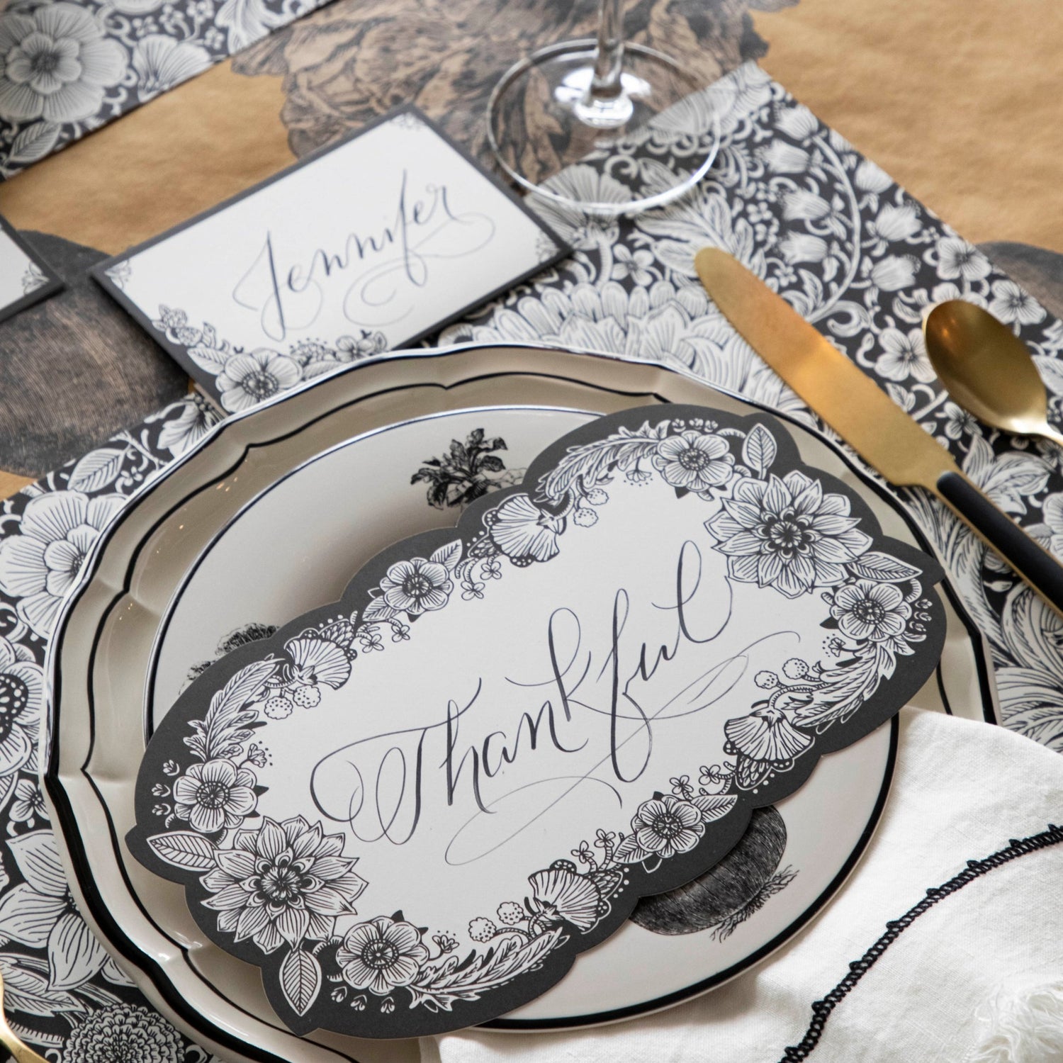 Close-up of an elegant place setting featuring an Ebony Harvest Blooms Table Card with &quot;Thankful&quot; written on it in lovely script resting on the plate.
