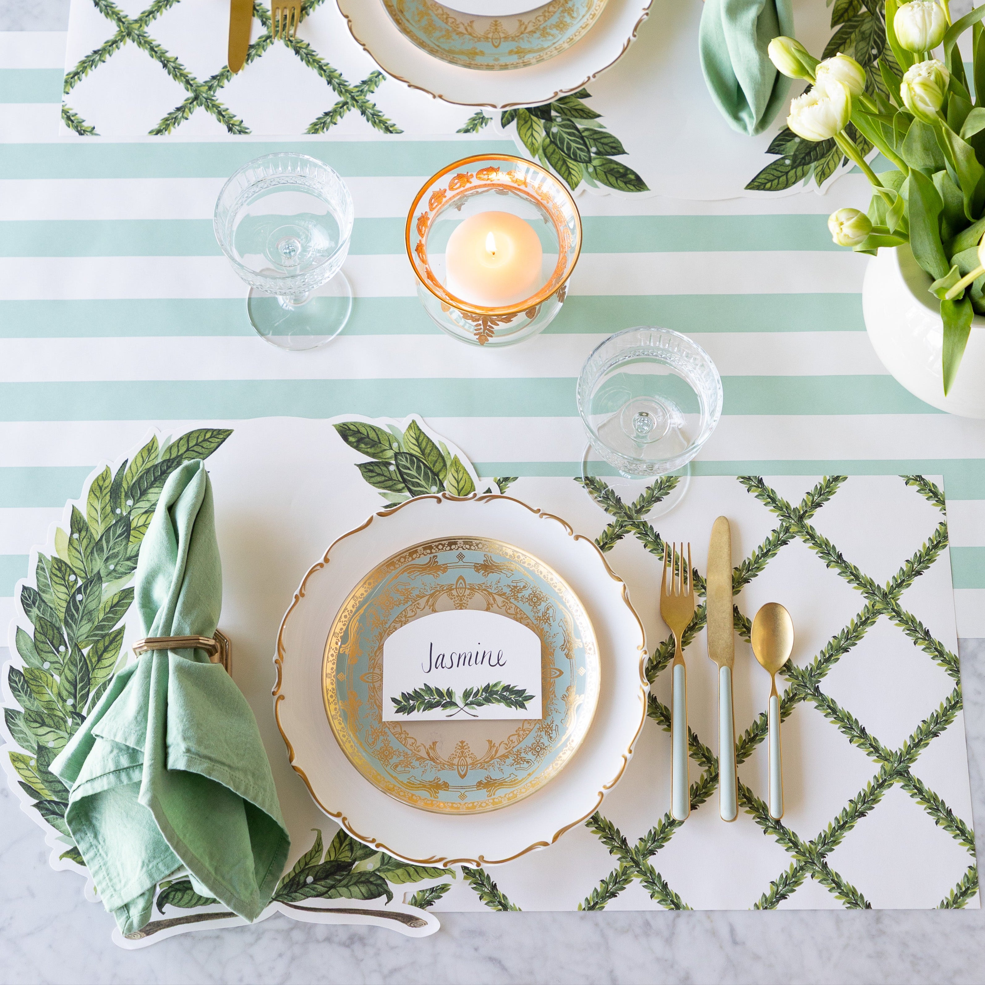 A table setting with a decorative plate, gold utensils, a green napkin in a ring, a glass, and candle sits atop the Die-cut Laurel Placemat and Laurel Lattice Placemat, with the Laurel place card labeled "Jasmine", on the Seafoam Classic Stripe Runner.