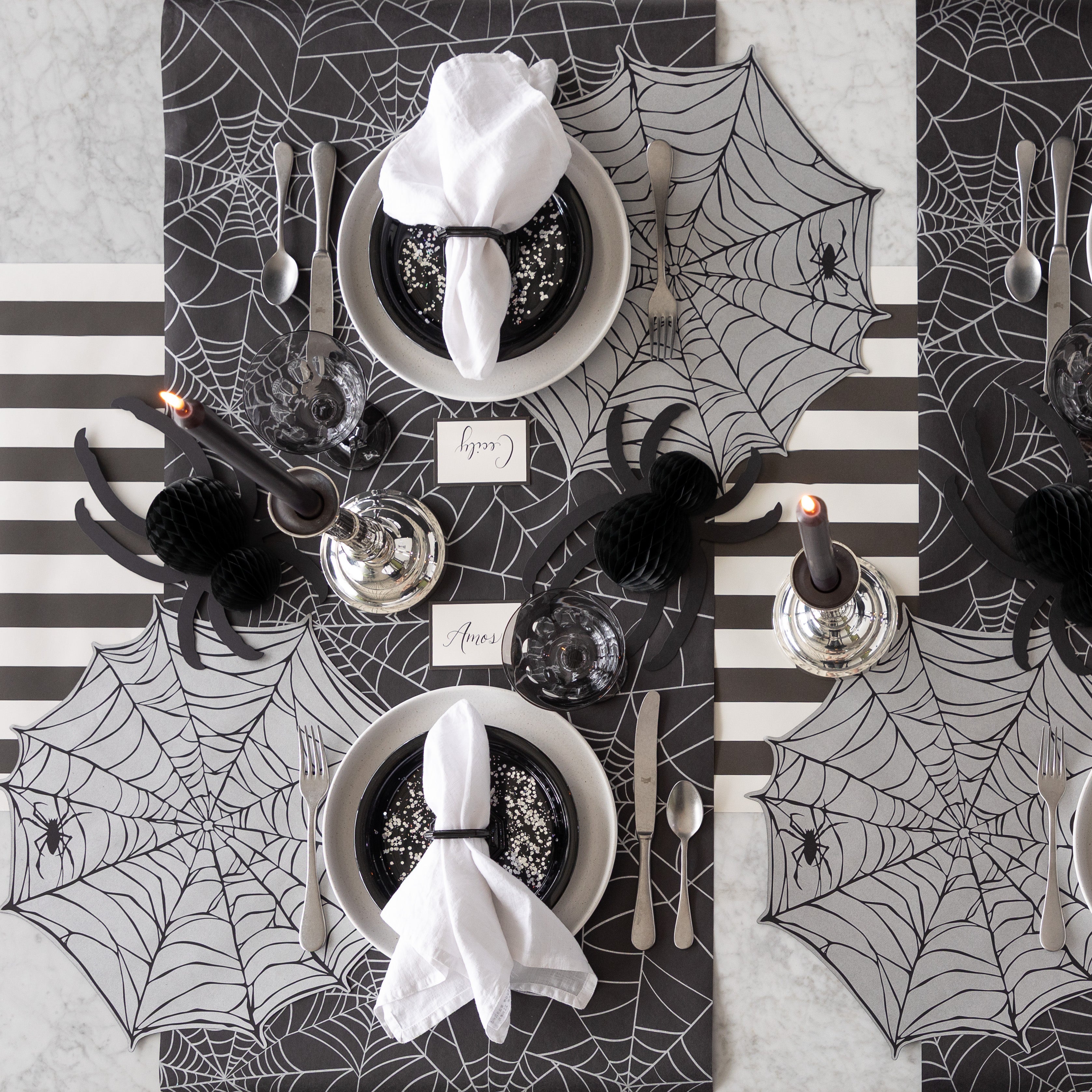 A Halloween-themed table setting with Die-cut Spiderweb Placemats, Honeycomb Spiders and Spiderweb and Black Classic Stripe Runner underneath.