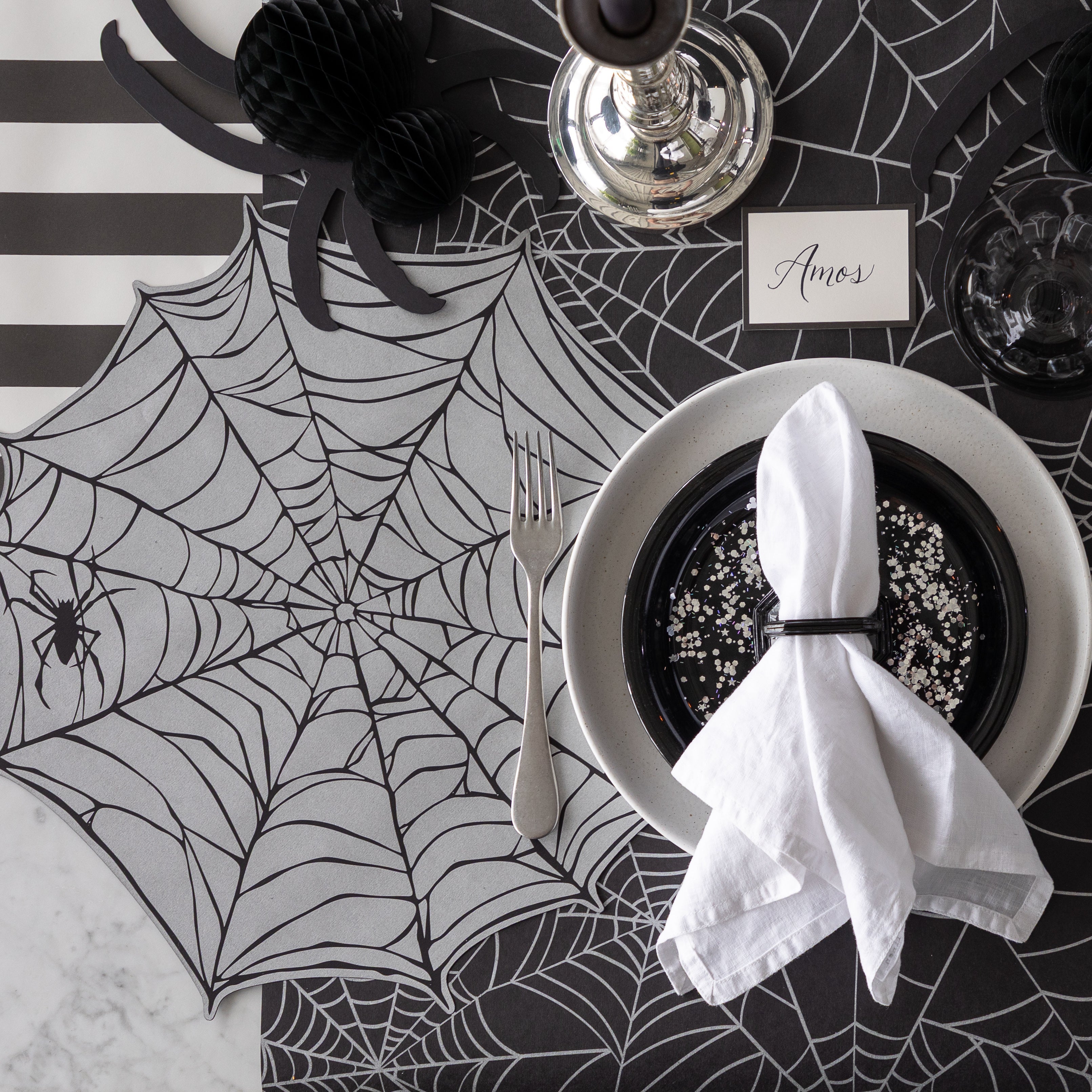A Halloween-themed place setting with the Die-cut Spiderweb Placemat, a honeycomb spider and Black Frame Place Card reading "Amos" on the Spiderweb Runner.