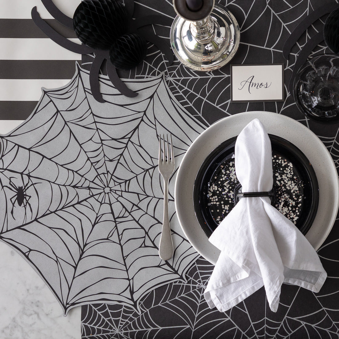 A Halloween-themed place setting with the Die-cut Spiderweb Placemat, a honeycomb spider and Black Frame Place Card reading "Amos" on the Spiderweb Runner.