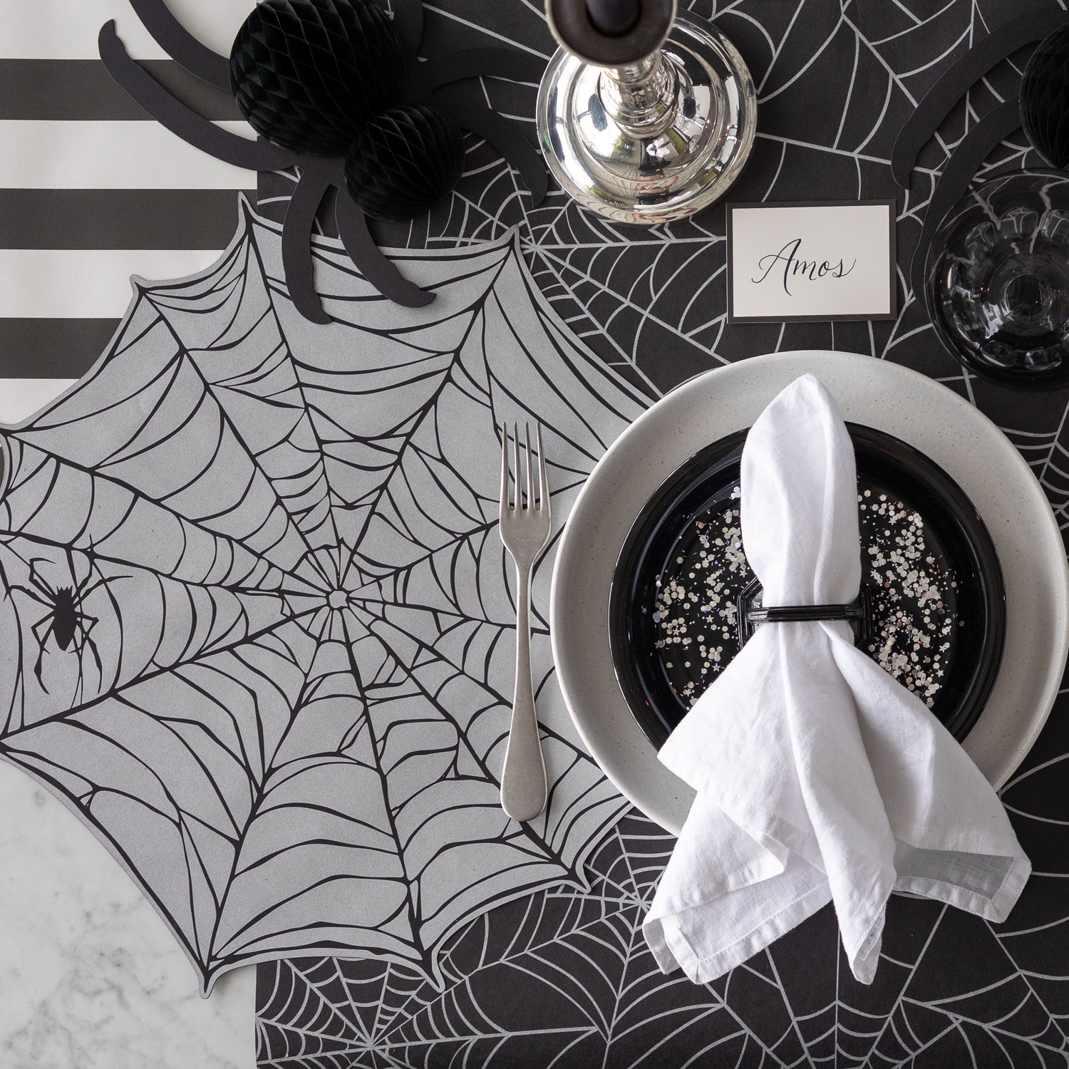 A Halloween-themed place setting with the Die-cut Spiderweb Placemat, a honeycomb spider and Black Frame Place Card reading &quot;Amos&quot; on the Spiderweb Runner.