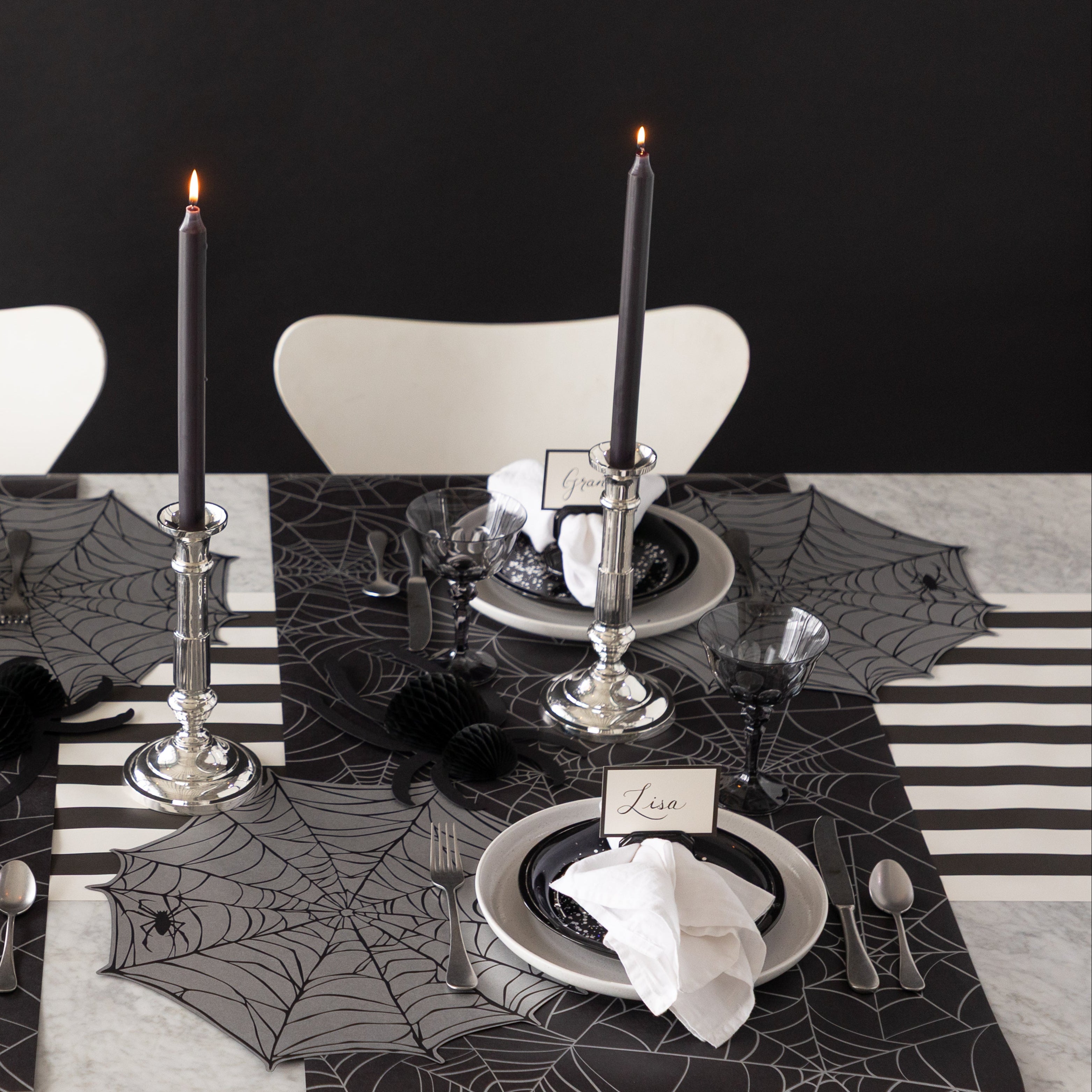 A Halloween-themed table setting featuring Die-cut Spiderweb Placemats, Spider Honeycomb and Black Frame Place Cards held by the Napkin Ring with Place Card Holder with Spiderweb Runner and Black Classic Stripe Runner underneath.