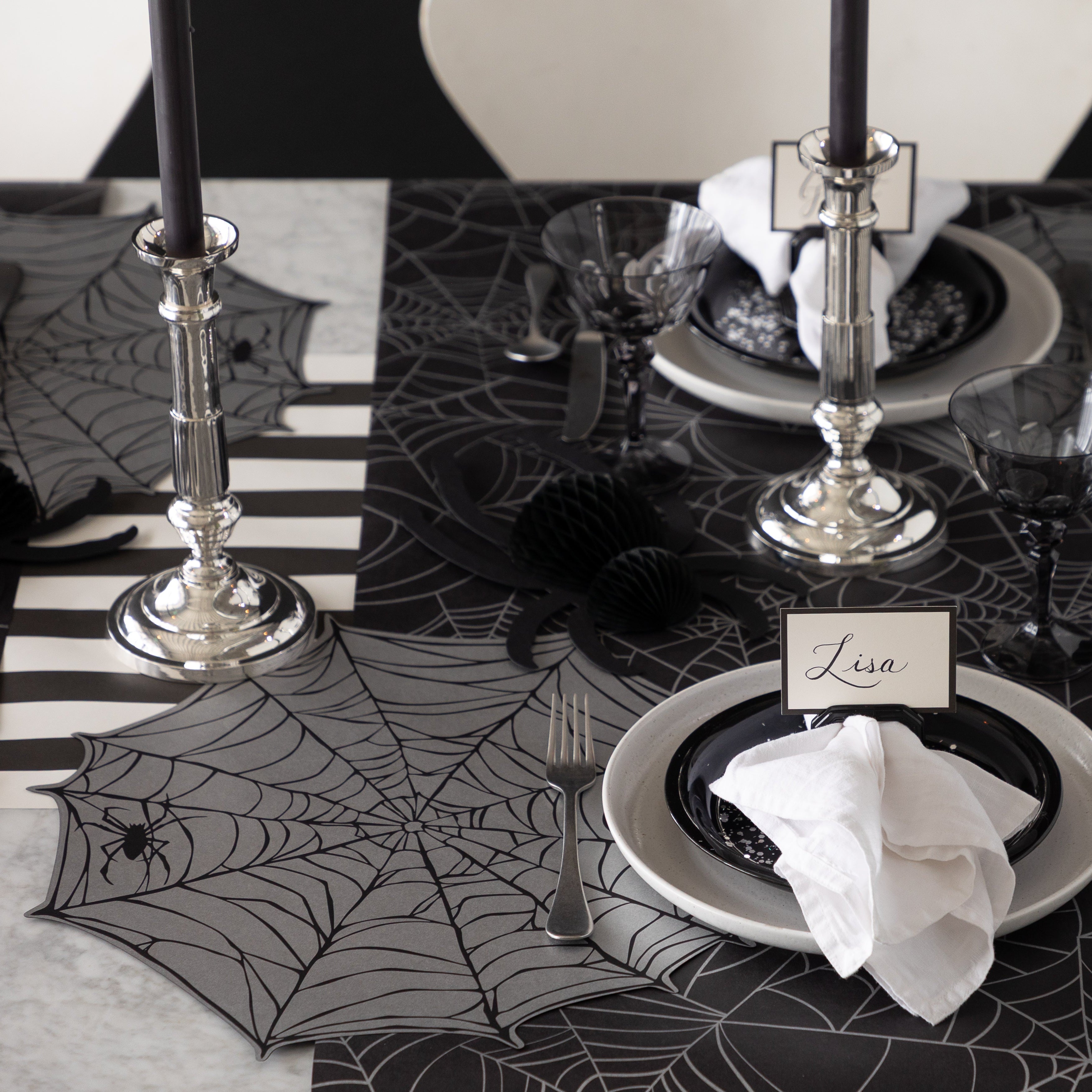 A Halloween-themed table setting featuring Die-cut Spiderweb Placemats, Spider Honeycomb and Black Frame Place Cards held by the Napkin Ring with Place Card Holder with Spiderweb Runner and Black Classic Stripe Runner underneath.