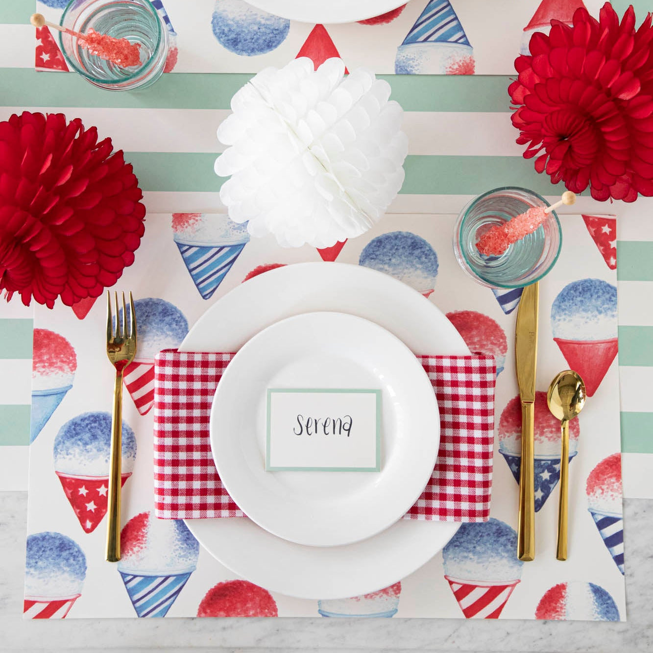 A patriotic place setting featuring the Snow Cone Placemat with white plates, a red-and-white checkered napkin, Seafoam Place Card reading &quot;Serena&quot; and gold flatware atop, on the Seafoam Classic Stripe Runner.