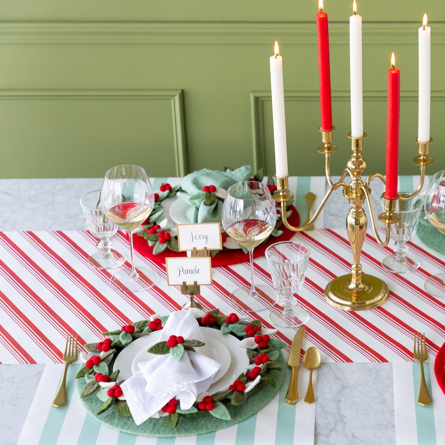A festive dining table set with Mint Felt Placemats, Felt Berry Chargers, Felt Berry Napkin Rings, gold cutlery, Gold Frame Place Cards, wine glasses, and red and white candles in a gold candelabra on the Peppermint Stripe and Seafoam Classic Stripe Runner.