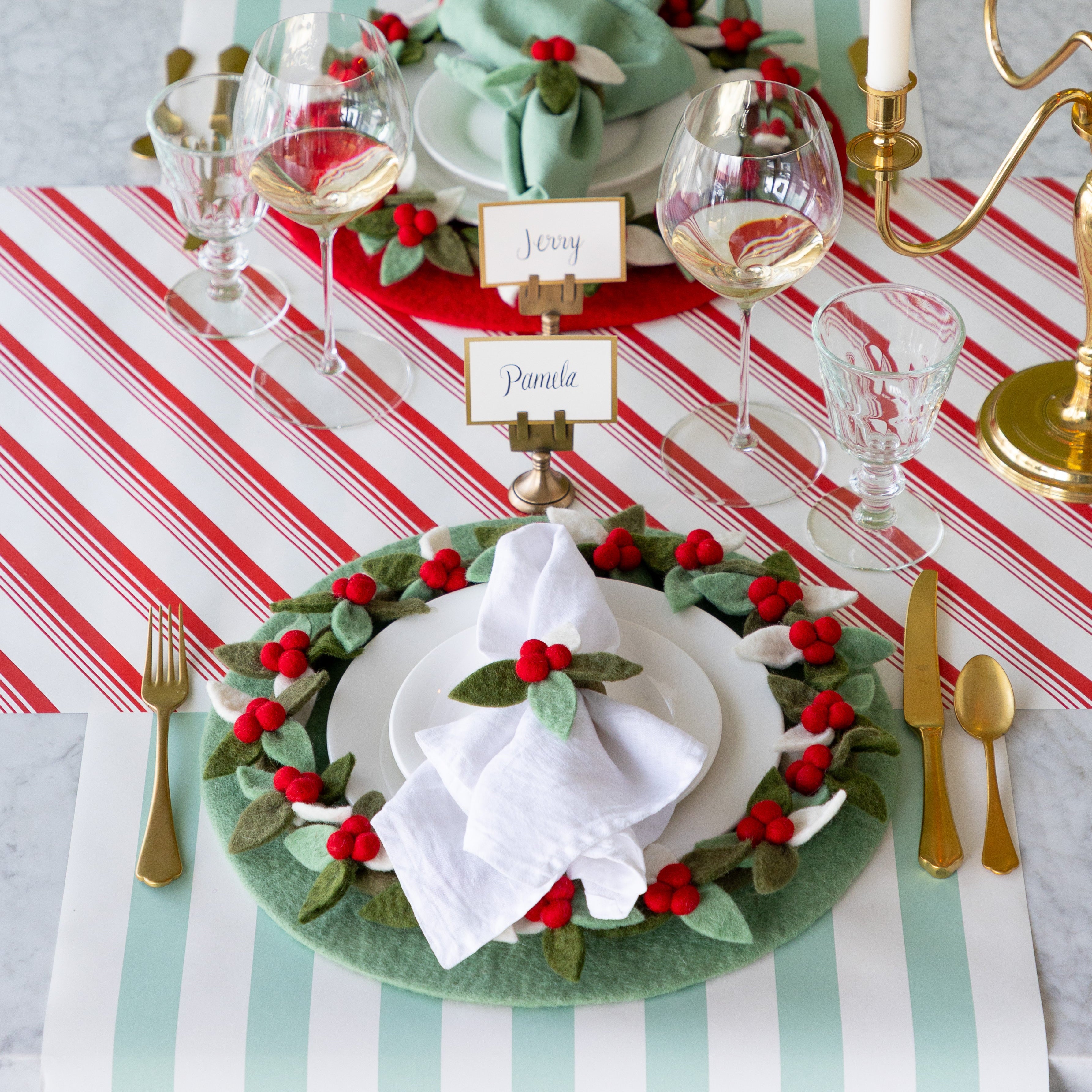 A festive dining table set with the Mint and Red Felt Placemats with napkins tied in the Felt Berry Napkin Rings and Felt Berry Wreath Charger atop, Gold Frame Place Cards, and gold cutlery on the Peppermint Stripe Runner and Seafoam Classic Stripe Runner.